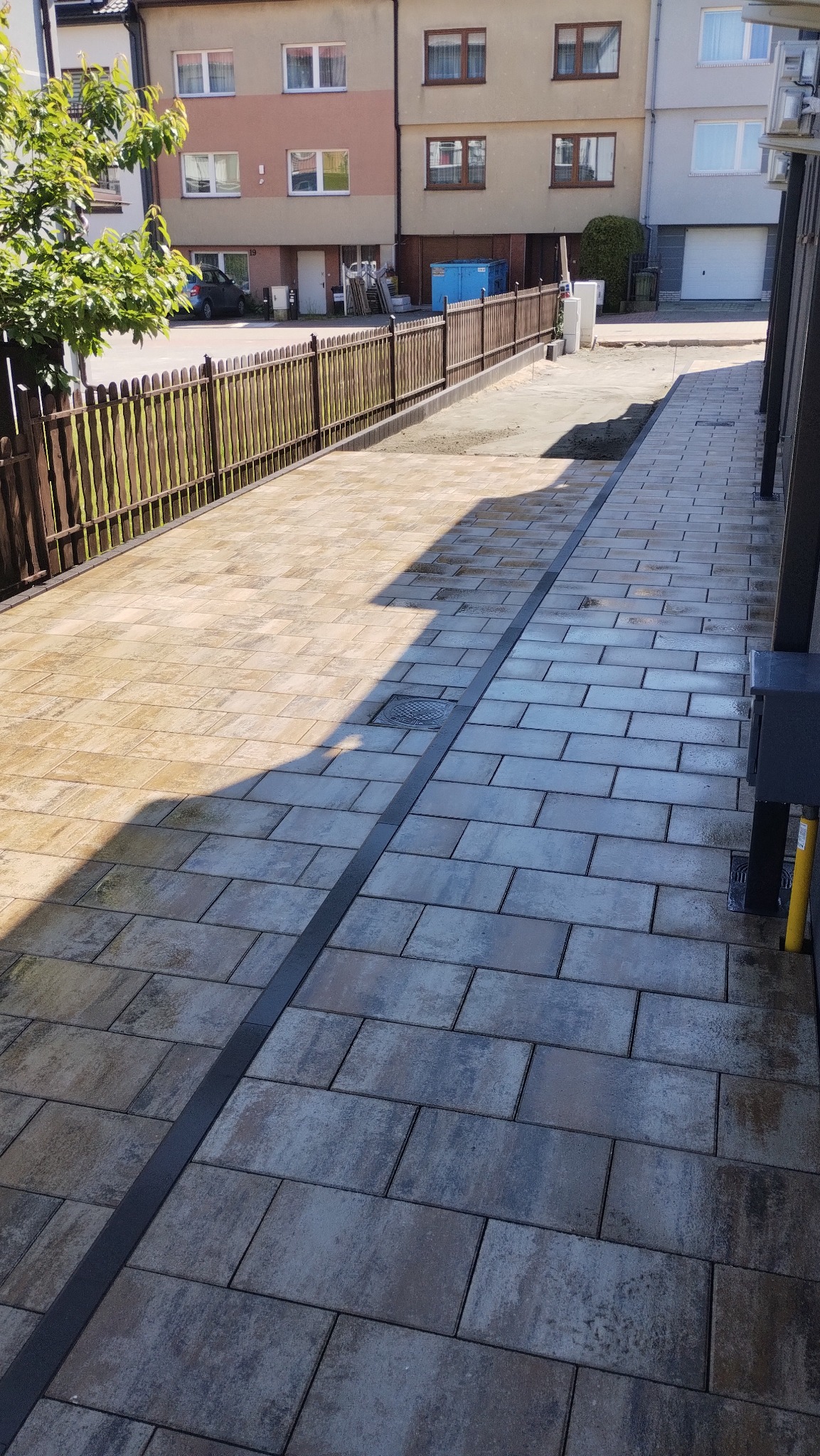 Ułożona kostka brukowa w odcieniach szarości i beżu z czarnym odpływem liniowym, prowadząca wzdłuż ogrodzenia i budynków mieszkalnych. Widoczne detale wykończenia.