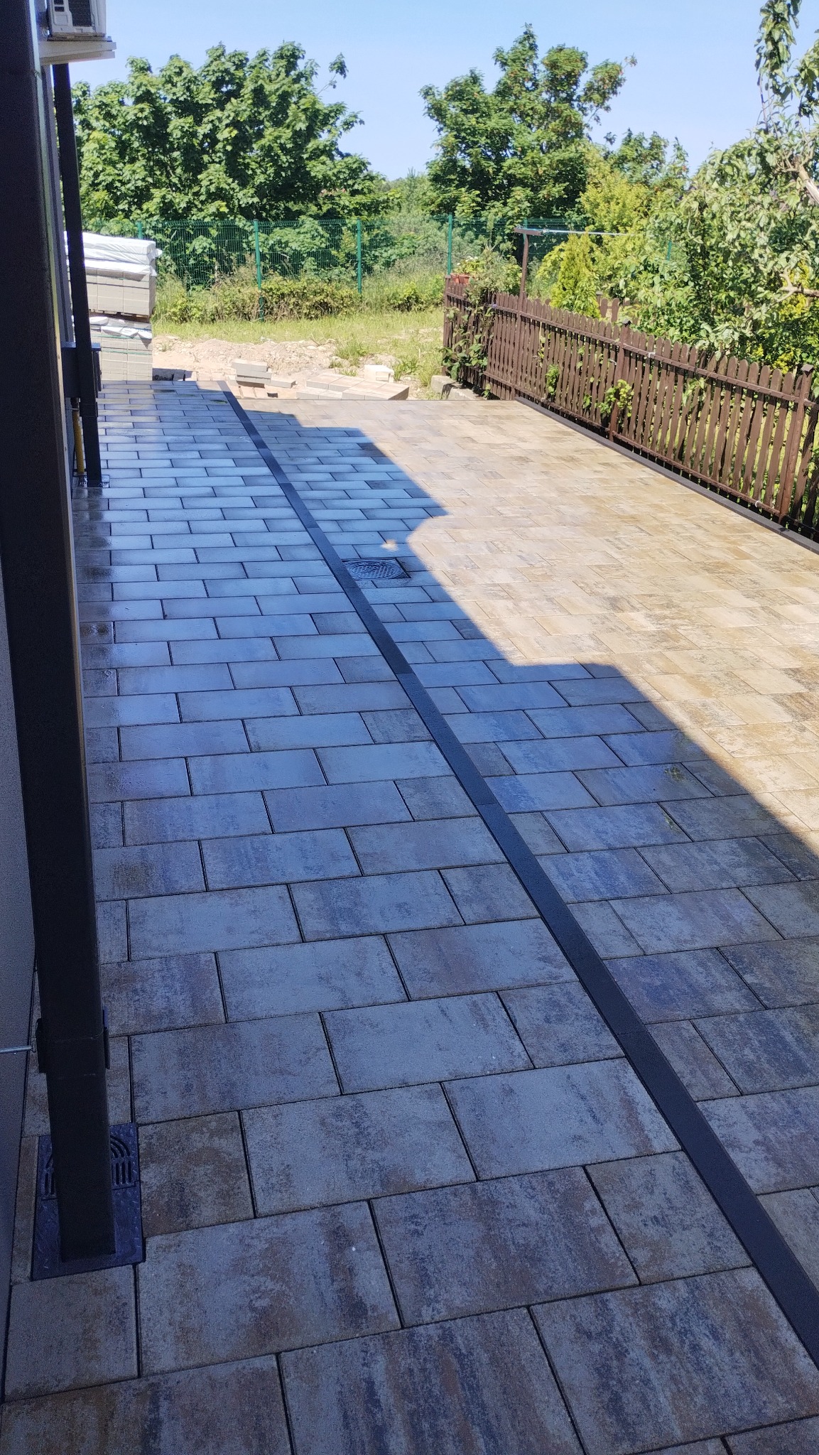 Ułożona kostka brukowa w odcieniach szarości i beżu z odpływem liniowym na posesji, widok na ogród z drewnianym płotem w słoneczny dzień.