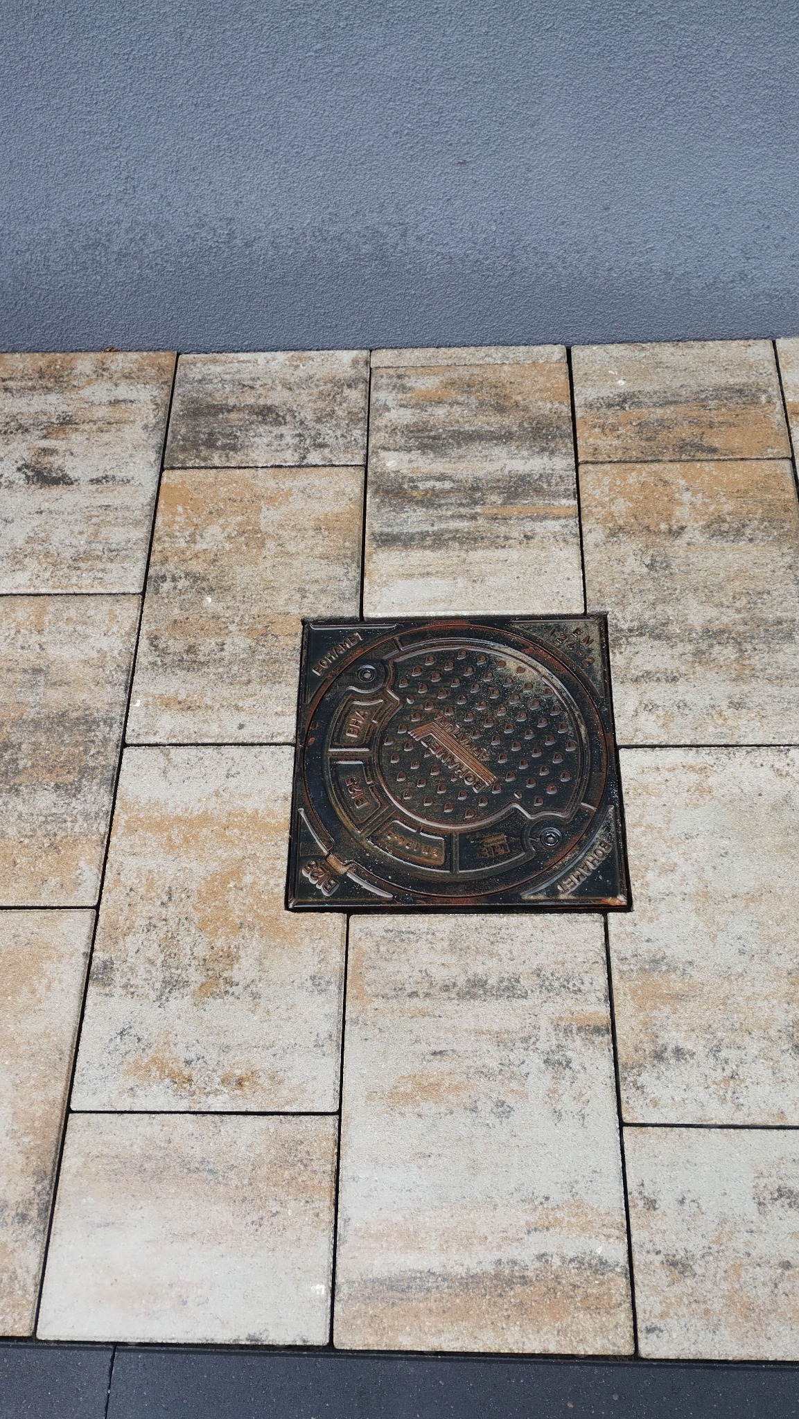 Ułożona kostka brukowa w Domatowie z włazem kanalizacyjnym. Beżowo-szare kostki tworzą wzór, a właz jest centralnym punktem. Tło stanowi szara ściana.