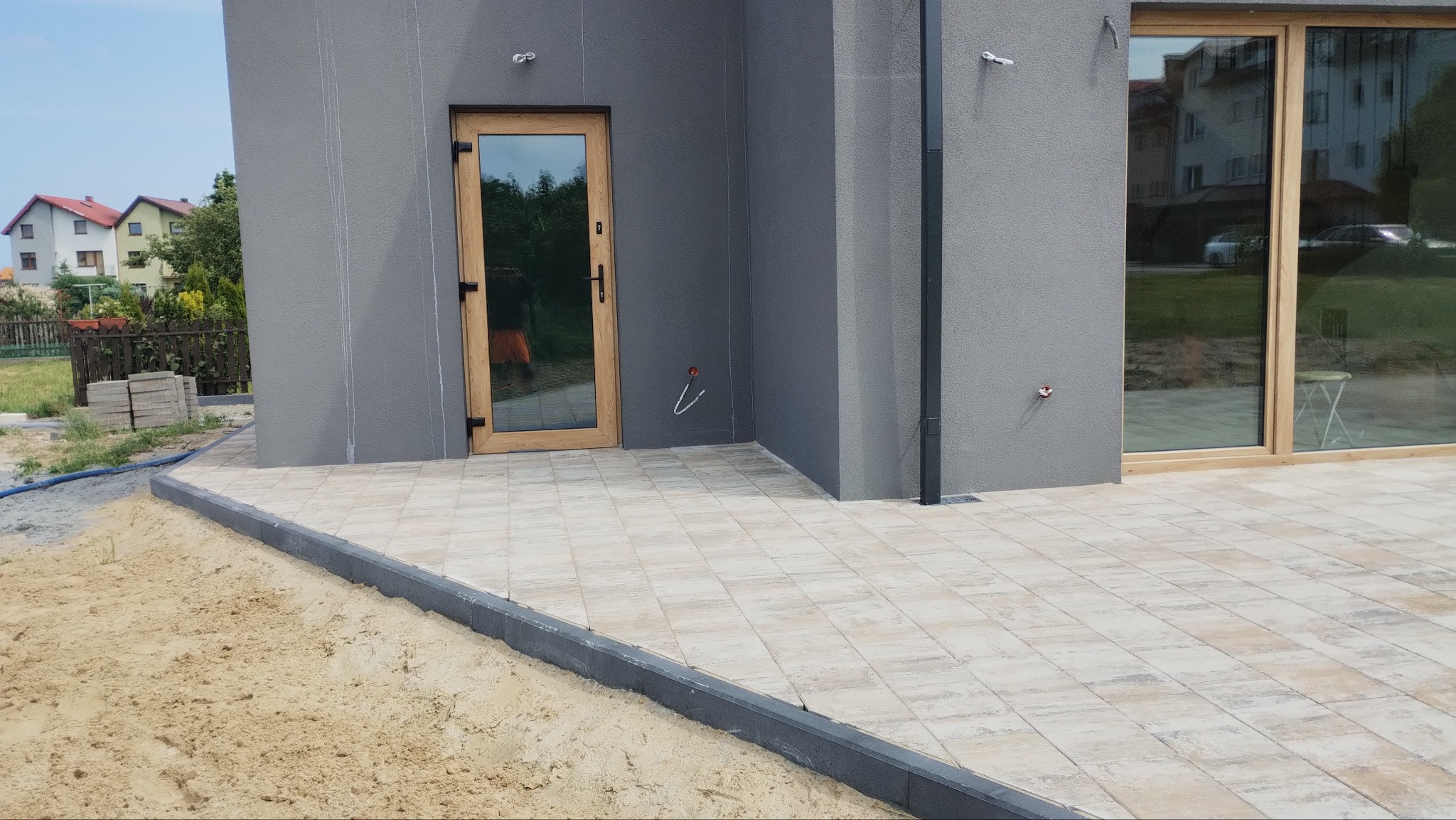 Ułożona jasna kostka brukowa przed szarym budynkiem z drewnianą stolarką okienną i drzwiową. Widoczne obrzeża i piasek. Częściowo ułożona kostka.