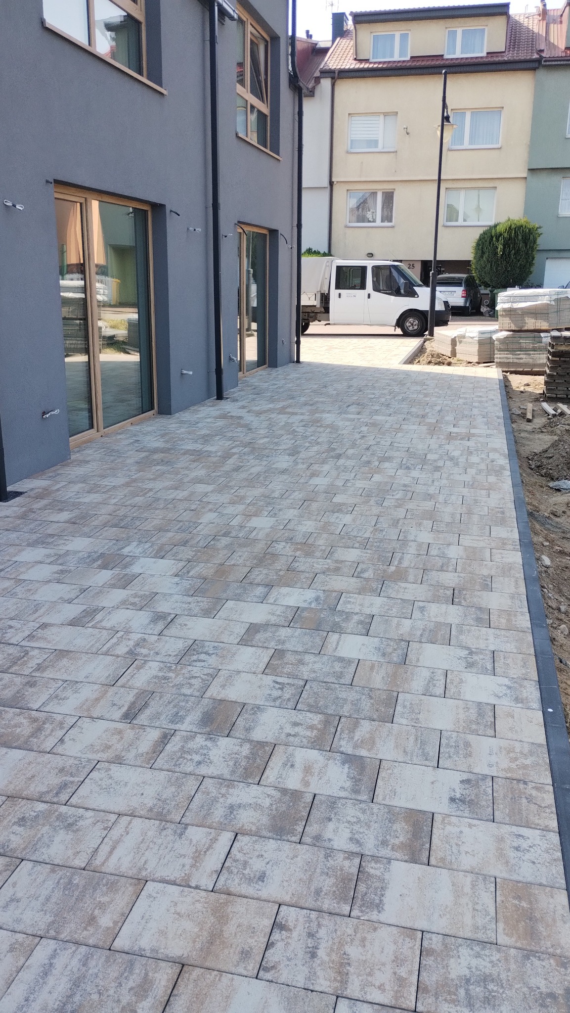 Ułożona kostka brukowa w odcieniach szarości i beżu przed budynkiem z szarymi ścianami i drewnianą stolarką okienną. W tle zabudowa miejska i zaparkowany biały bus.