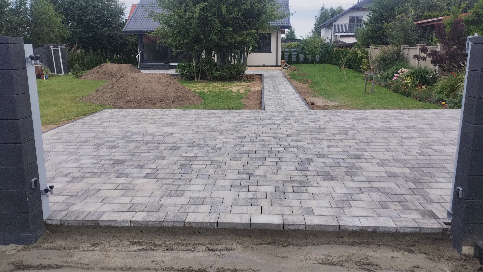 Ułożona kostka brukowa w odcieniach szarości tworzy duży podjazd przed domem. Widoczne bramy wjazdowe i ścieżka prowadząca do budynku. Na trawniku usypana ziemia.