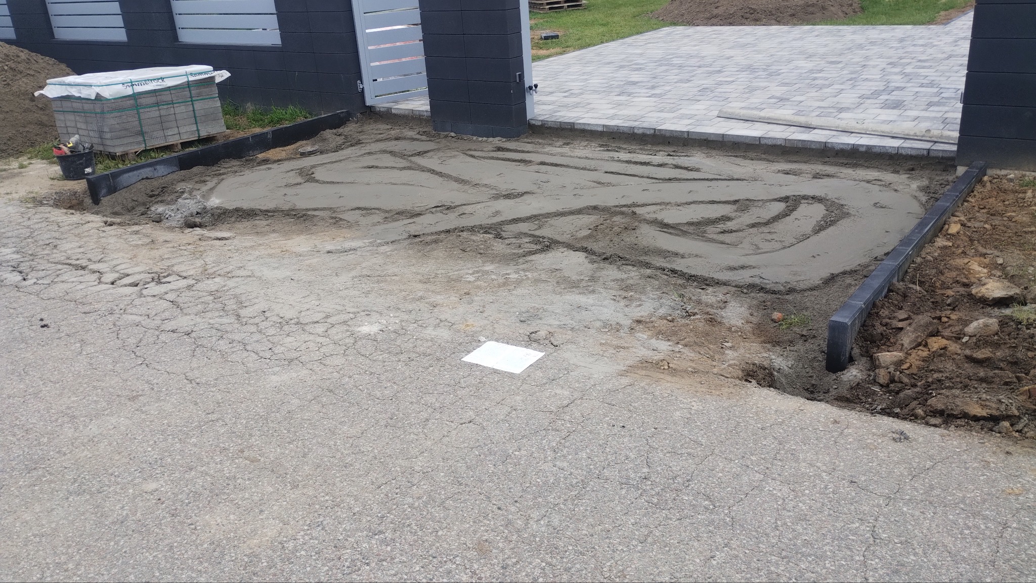 Przygotowanie podłoża pod kostkę brukową: wyrównany teren z obrzeżami, widoczny zapas kostki, brama w tle. Etap prac przed ułożeniem kostki.