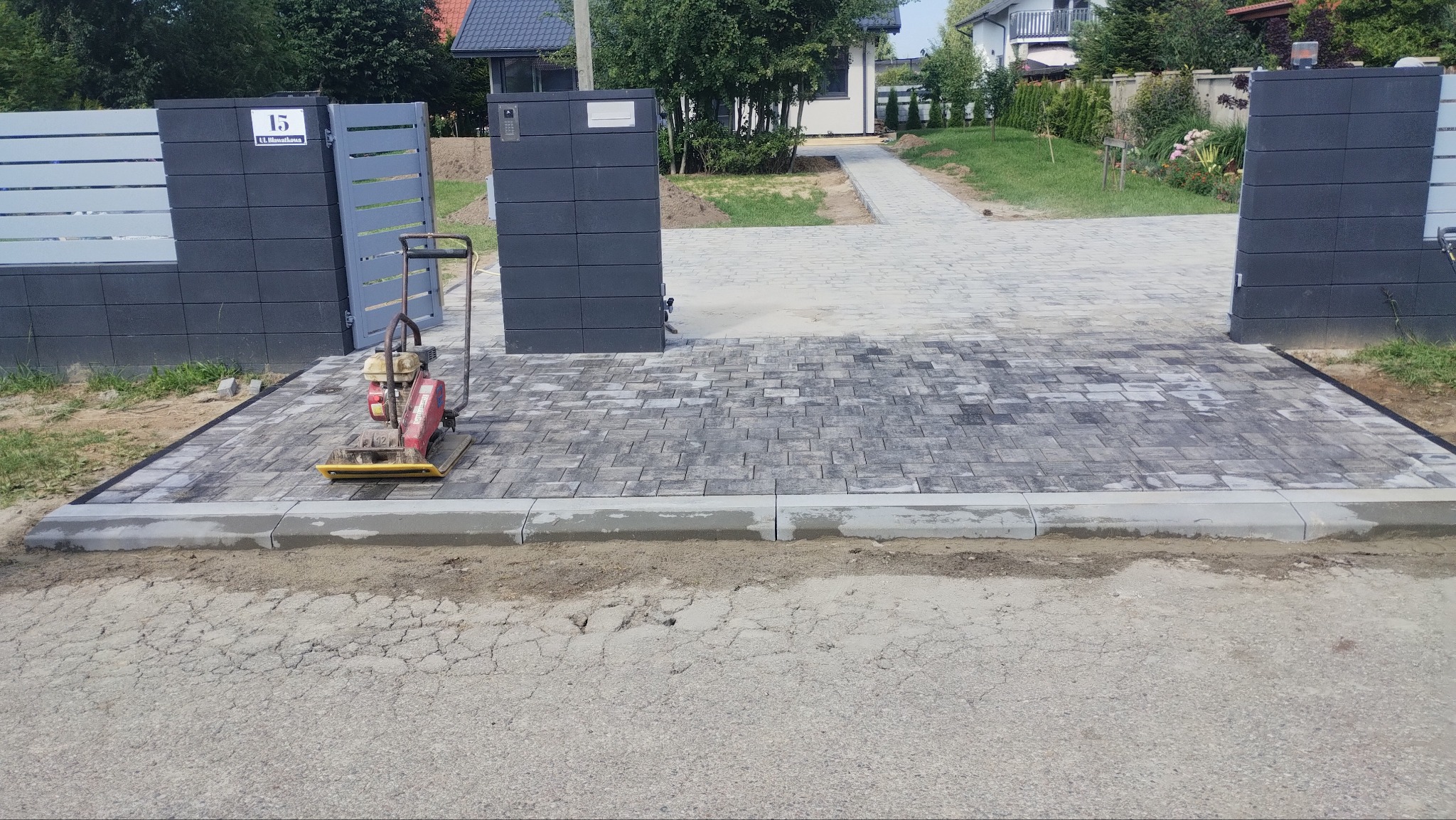Ułożona kostka brukowa w szaro-białej tonacji, z widoczną zagęszczarką w pobliżu nowoczesnego ogrodzenia posesji z numerem 15. Widoczny chodnik z kostki prowadzący do domu.