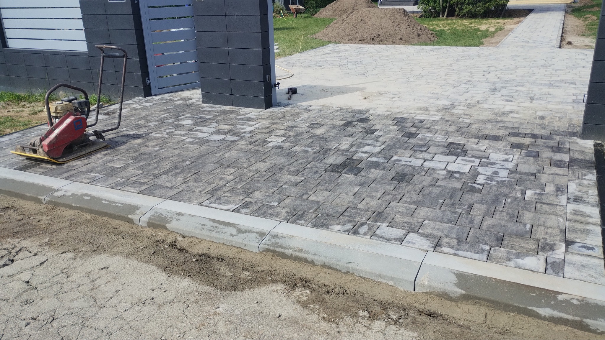 Ułożona kostka brukowa w odcieniach szarości, z widoczną ubijarką mechaniczną i krawężnikiem. W tle brama wjazdowa i hałda ziemi.