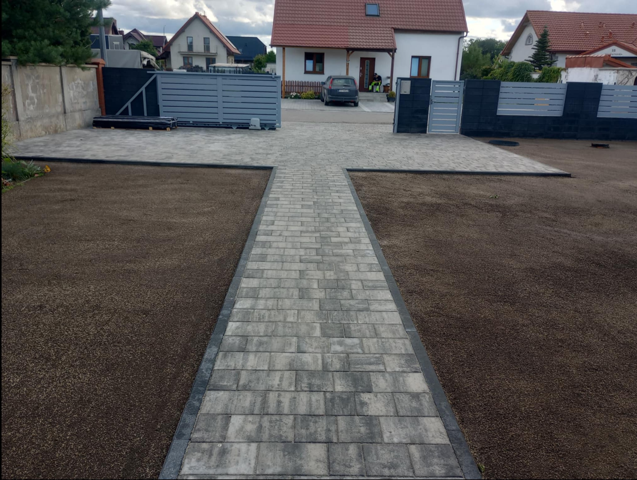 Ułożona kostka brukowa w kształcie krzyża prowadzi do domu z szarym ogrodzeniem i bramą. Świeżo posiana trawa na rabatach. Widok z perspektywy pieszego.