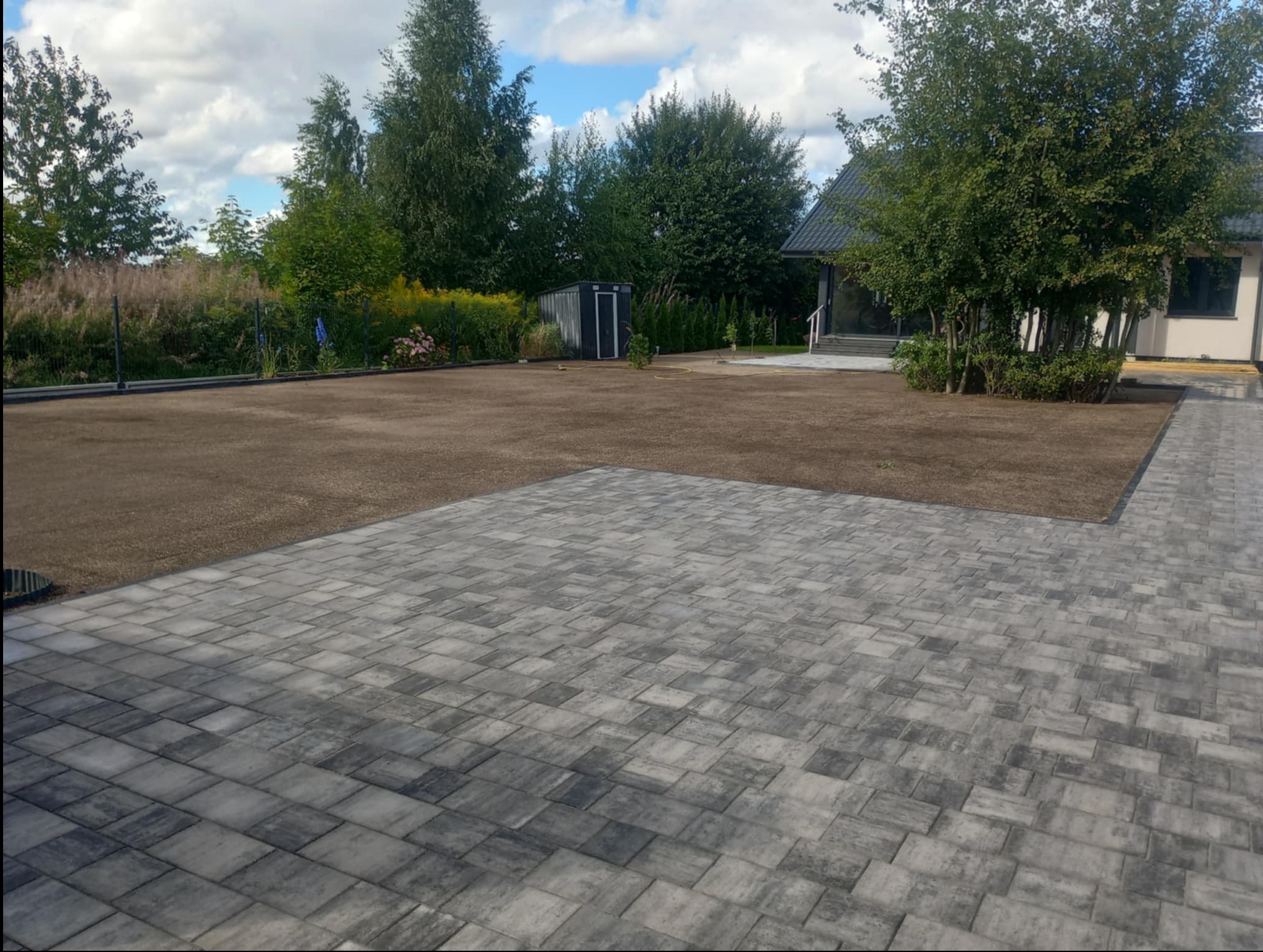 Nowo ułożona, szara kostka brukowa na podjeździe, kontrastująca z przygotowanym terenem pod trawnik i zielenią ogrodu w tle, w słoneczny dzień.