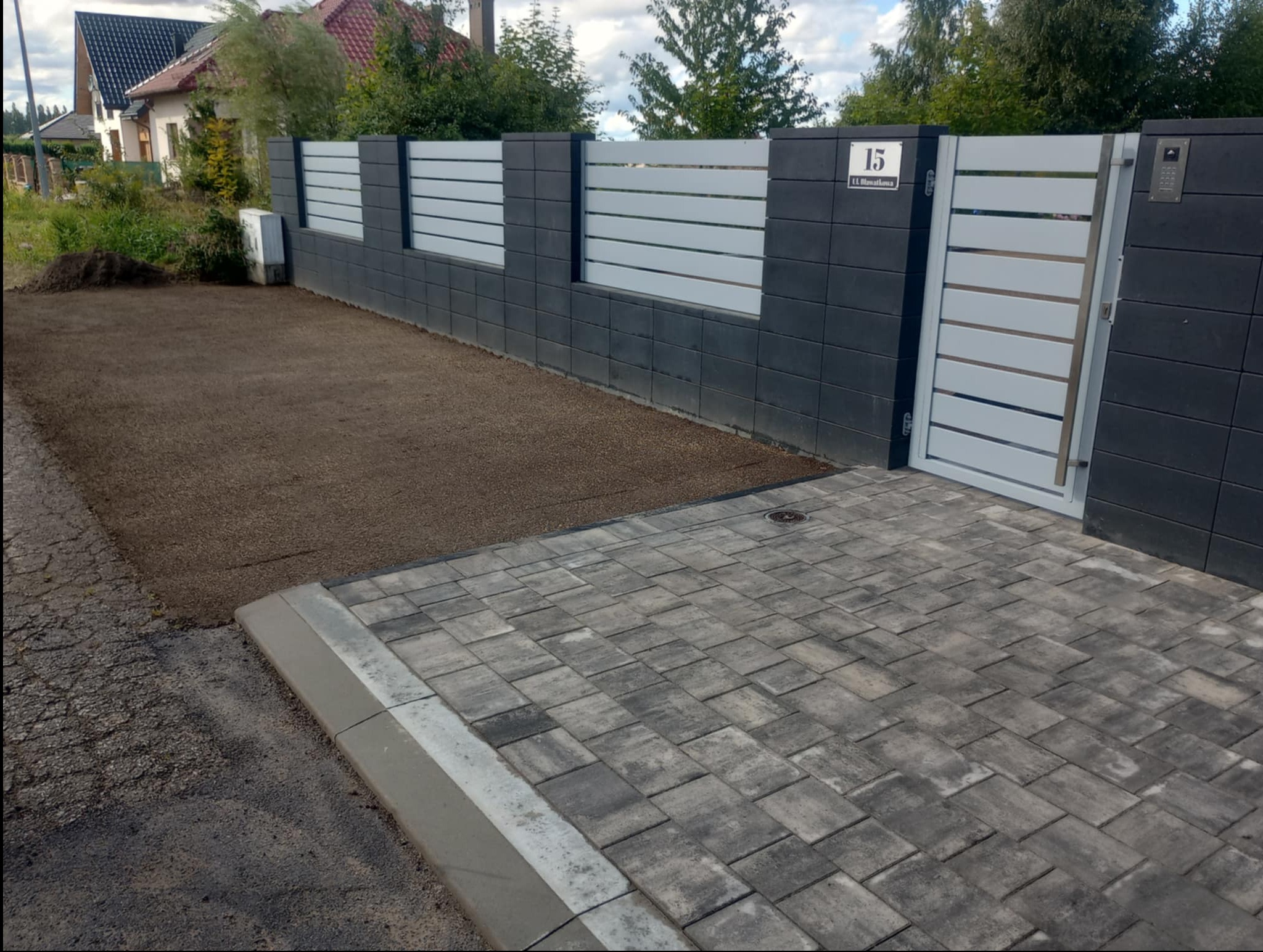 Nowoczesna posesja z szarej kostki brukowej i ogrodzeniem z numerem domu, z fragmentem podłoża pod trawnik. Widok z poziomu ulicy.