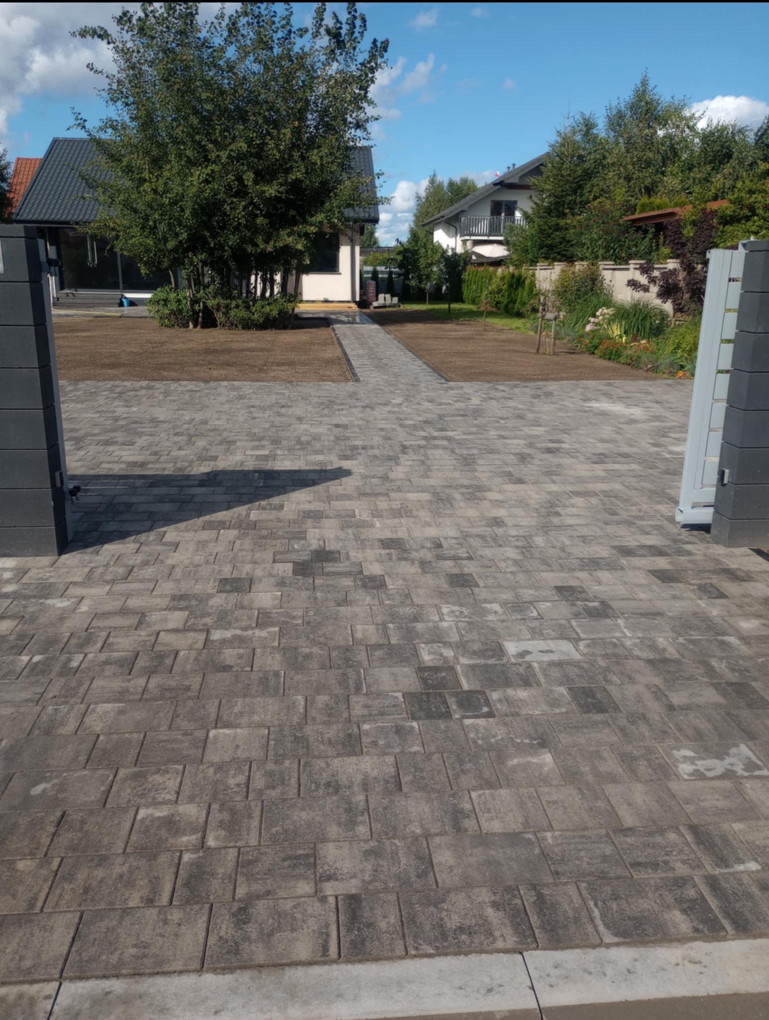 Szara kostka brukowa ułożona na podjeździe do domu, widok z poziomu gruntu, brama wjazdowa po bokach kadru, świeżo posiana trawa w tle.