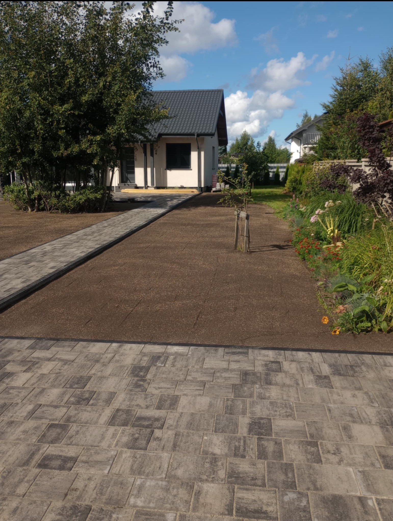 Ułożona kostka brukowa w odcieniach szarości tworzy ścieżkę i obrzeże. W tle dom z ciemnym dachem, trawnik i rabaty kwiatowe, wszystko w słoneczny dzień.