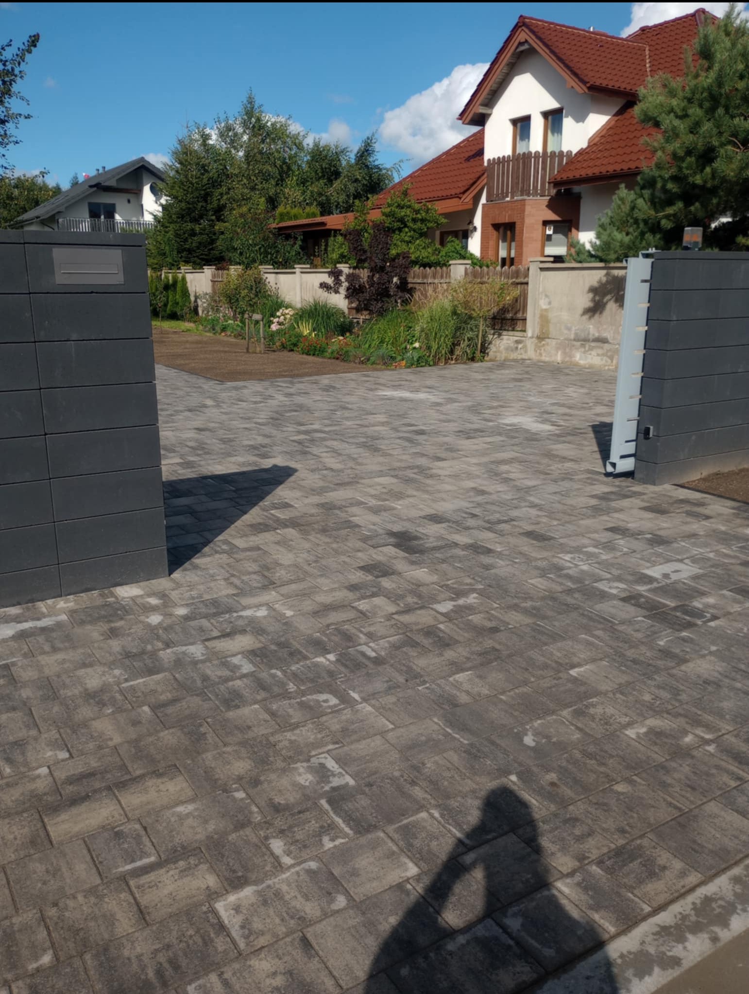 Ułożona kostka brukowa w odcieniach szarości prowadząca do otwartej bramy wjazdowej. W tle dom z czerwonym dachem i zadbany ogród.