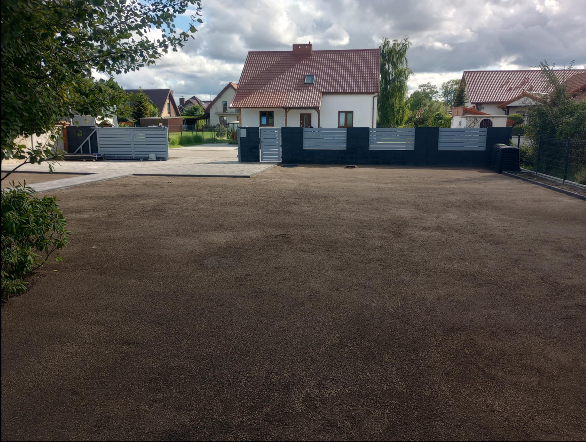 Przygotowany teren pod trawnik z widocznym ogrodzeniem panelowym i kostką brukową. W tle dom jednorodzinny i błękitne niebo z chmurami.