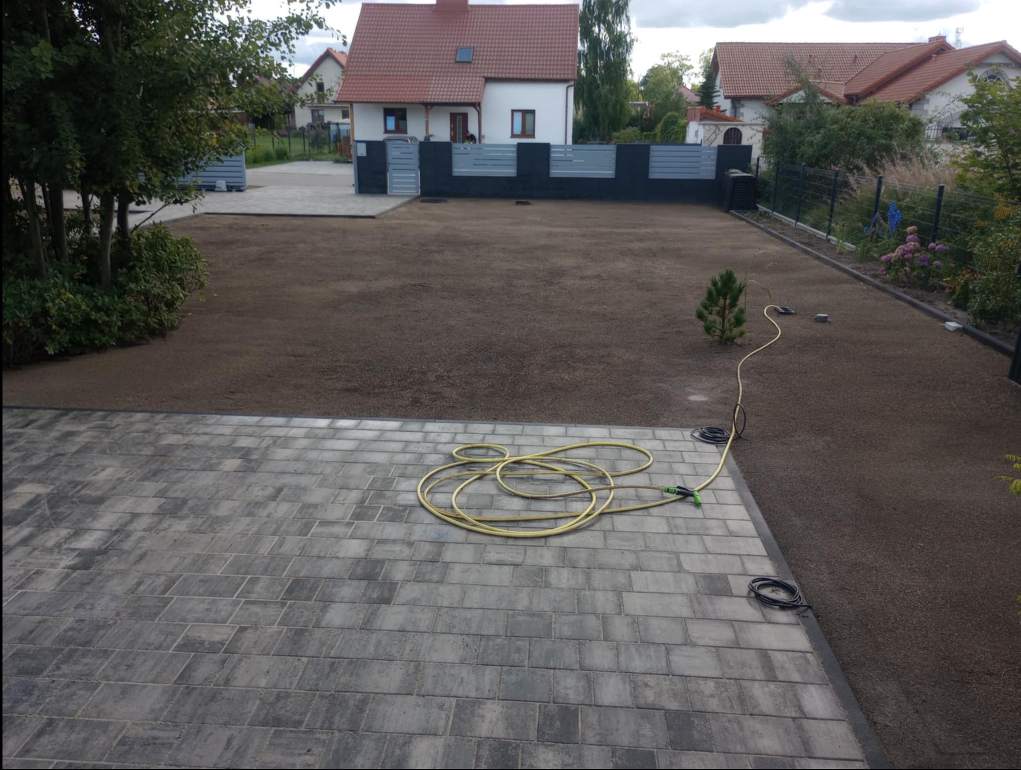 Ułożona kostka brukowa na podjeździe domu jednorodzinnego, z widocznym przygotowanym terenem pod trawnik i elementami ogrodzenia. Wąż ogrodowy na kostce.