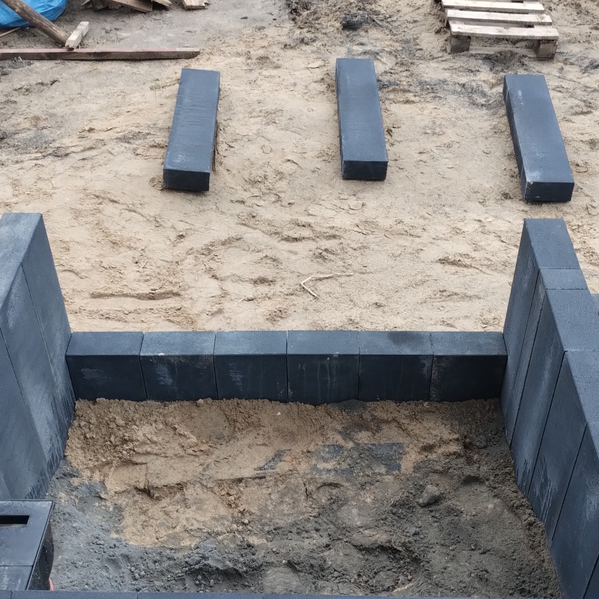 Etap budowy: czarne, prostokątne elementy obrzeża ułożone w piasku, tworzące ramę. Widoczne przygotowania do dalszych prac ziemnych.