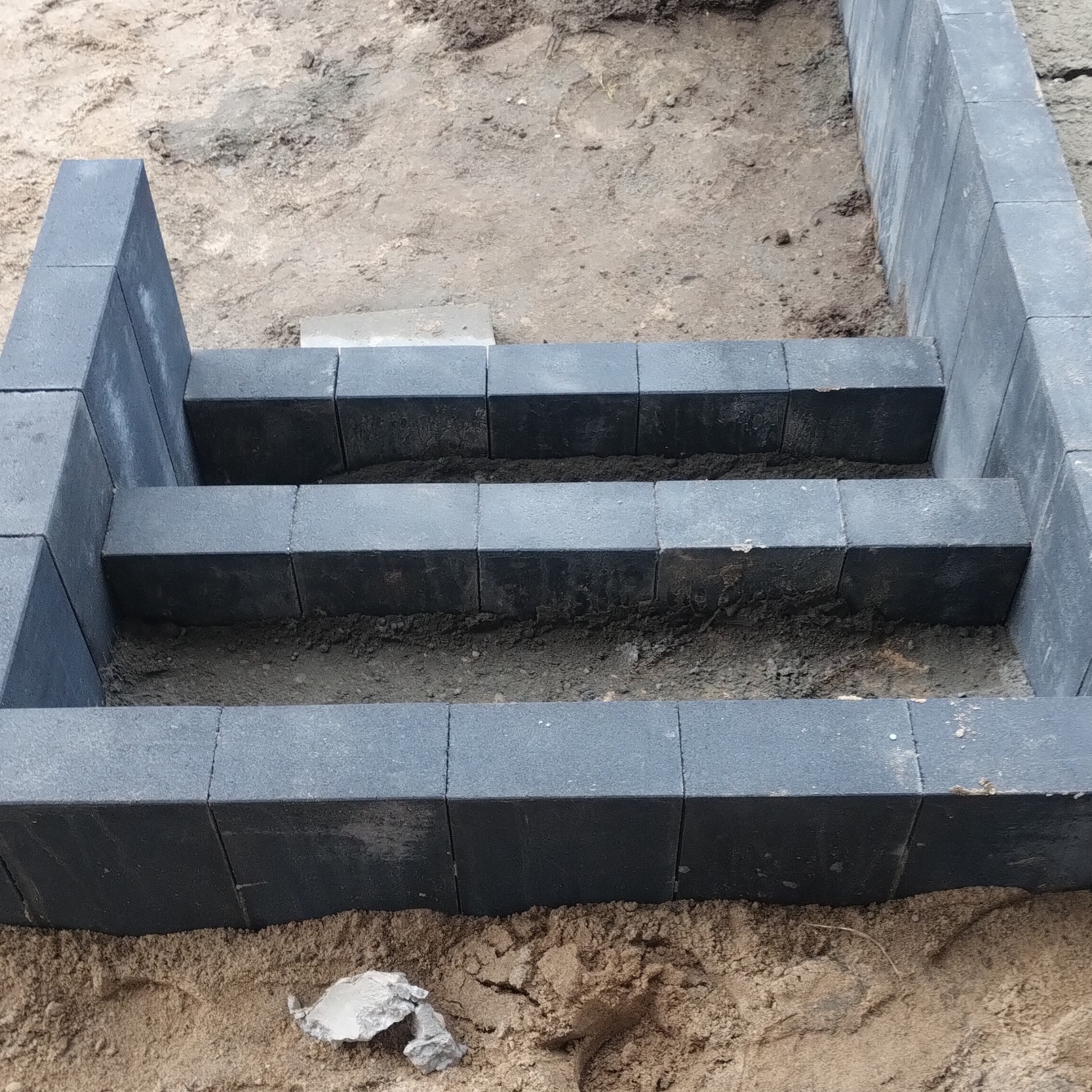 Schody z ciemnej kostki brukowej w trakcie budowy, widoczne warstwy piasku i ziemi. Ujęcie z góry, skupione na detalach konstrukcji.