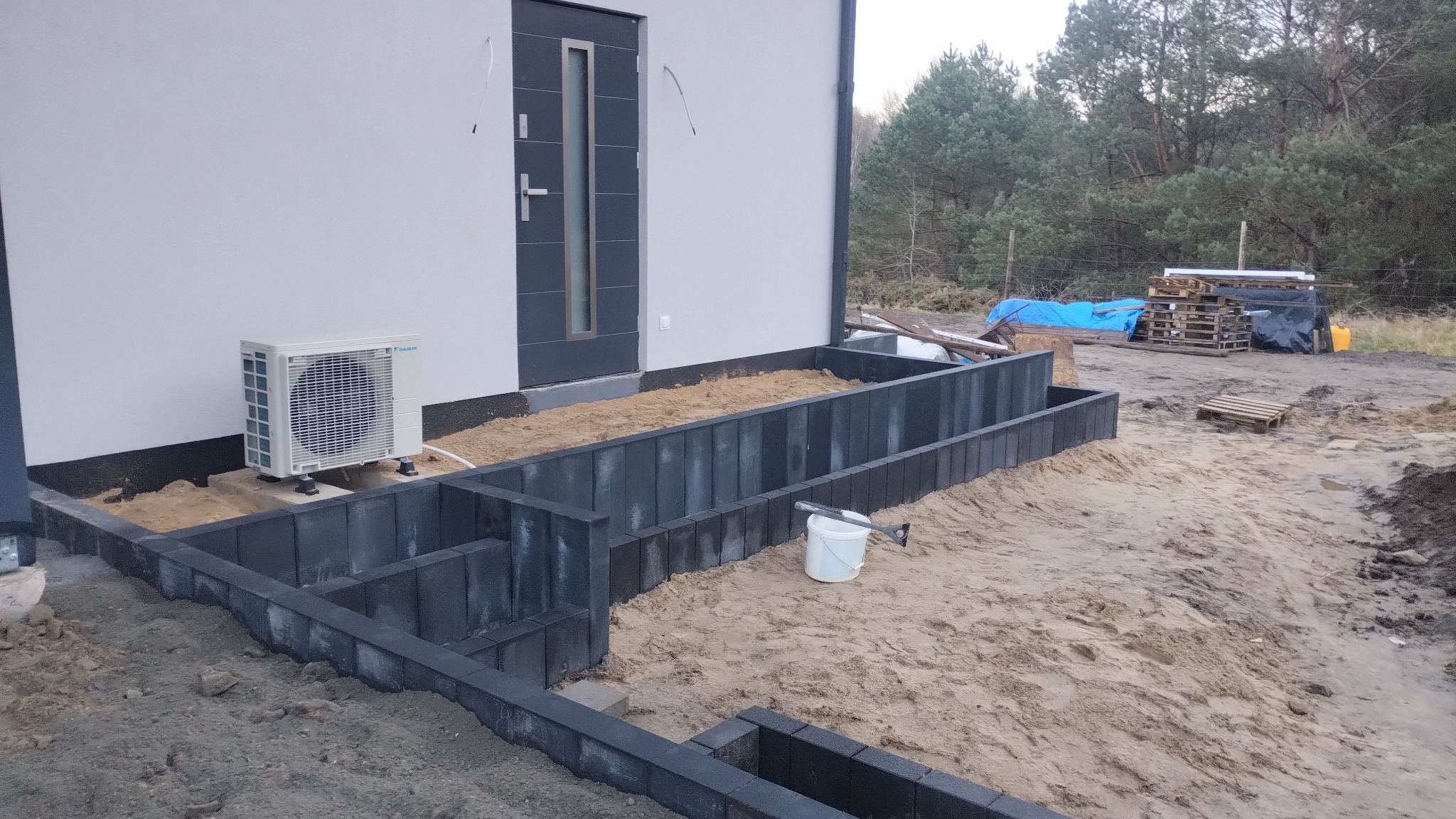 Ciemnoszara obudowa z kostki przy białym budynku, piasek wewnątrz, widoczny klimatyzator i narzędzia. Etap prac ziemnych przed dalszym wykończeniem.