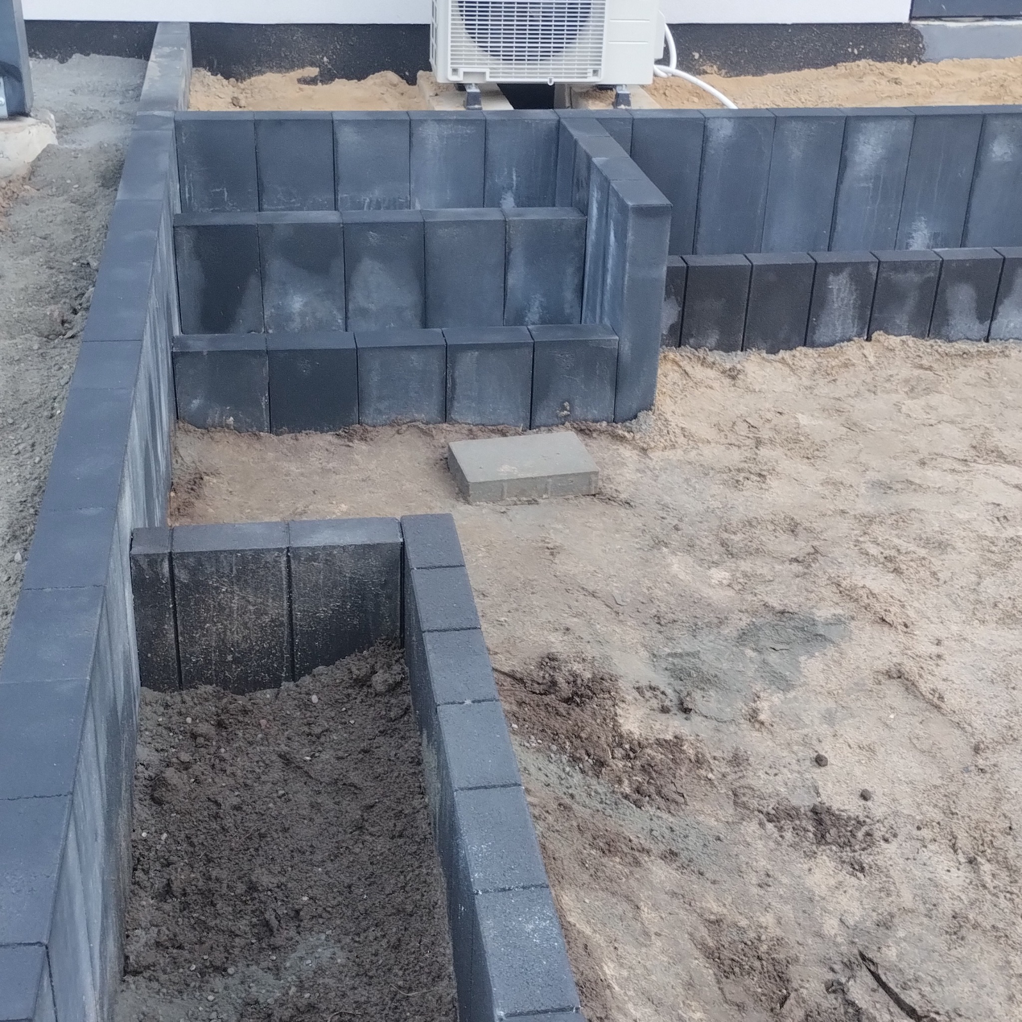 Częściowo ułożona, grafitowa kostka brukowa tworząca obrzeże rabaty na piasku. Widoczny klimatyzator na ścianie budynku w tle. Etap prac brukarskich.