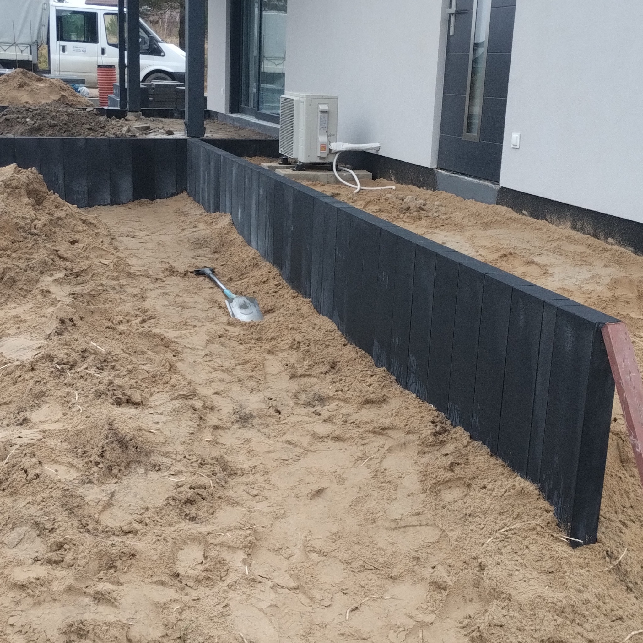 Montaż obrzeży trawnikowych z szarej kostki betonowej, widoczna łopata wbita w piasek, budynek w tle. Obrzeża oddzielają trawnik od elewacji.