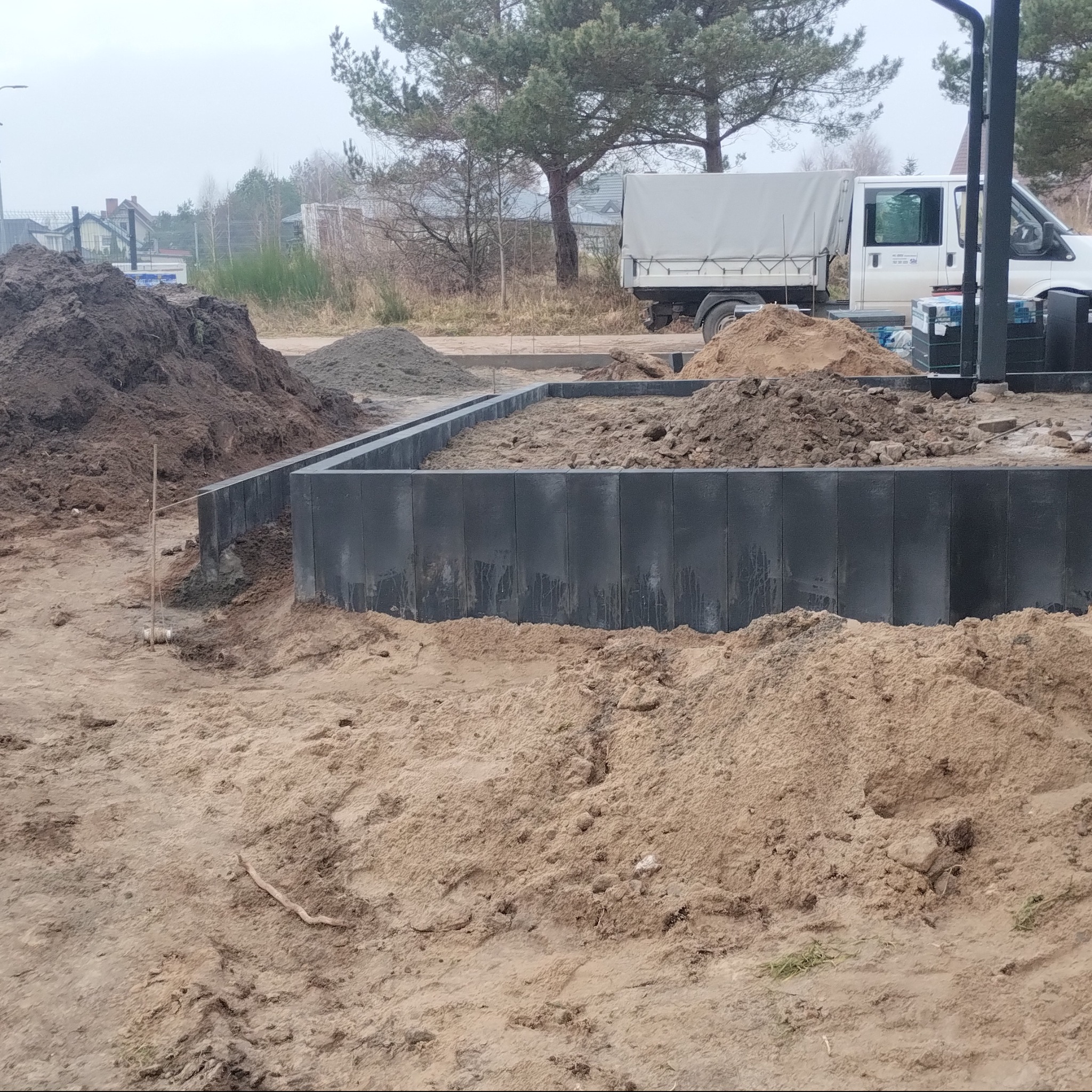 Częściowo ułożona, grafitowa obudowa rabaty kwiatowej z kostki. W tle sterta ziemi i piasku, oraz biały samochód dostawczy. Pochmurny dzień.