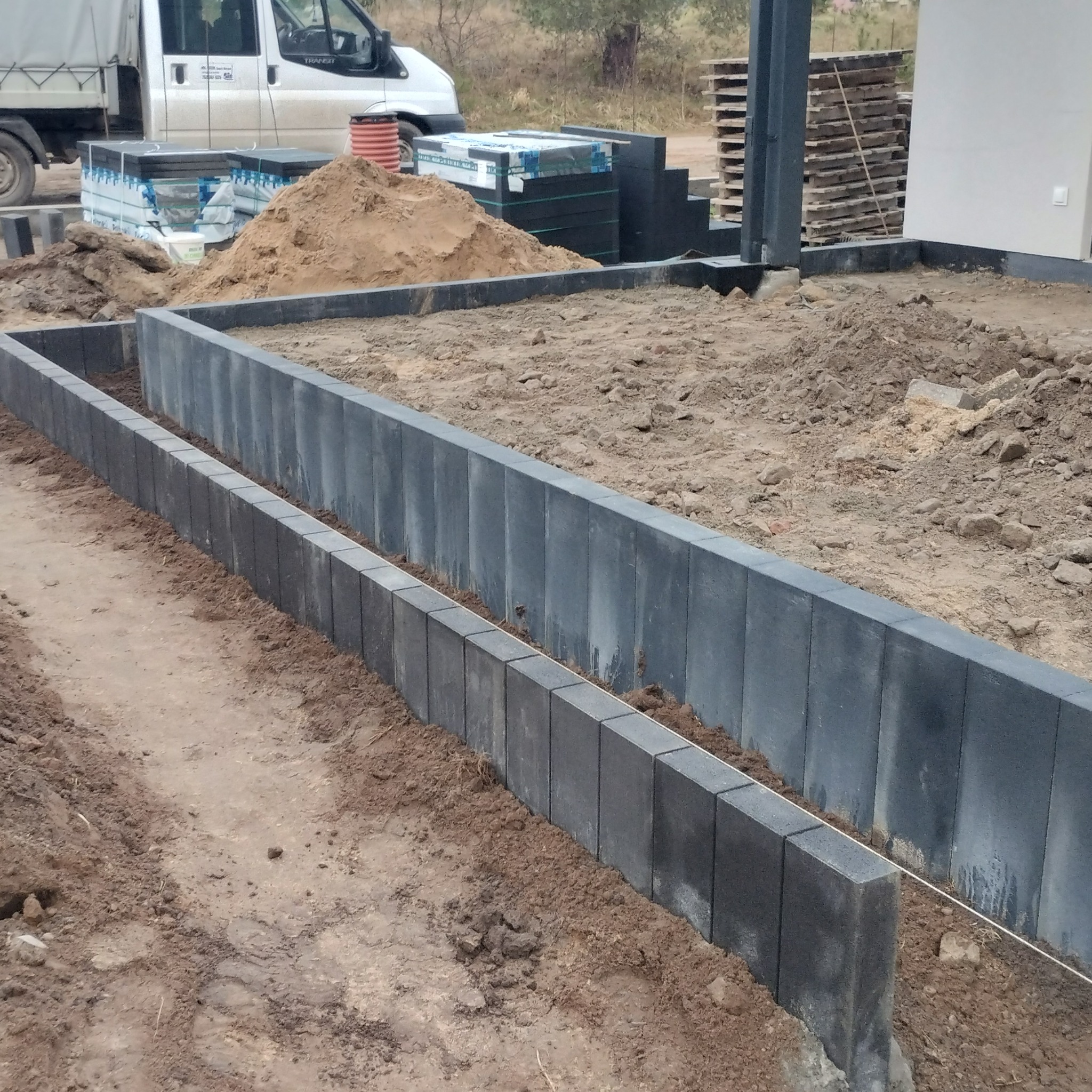 Wyrównane obrzeża z szarej kostki betonowej, wyznaczające obszar przyszłego chodnika. W tle sterta piasku, palety z kostką i biały samochód dostawczy.