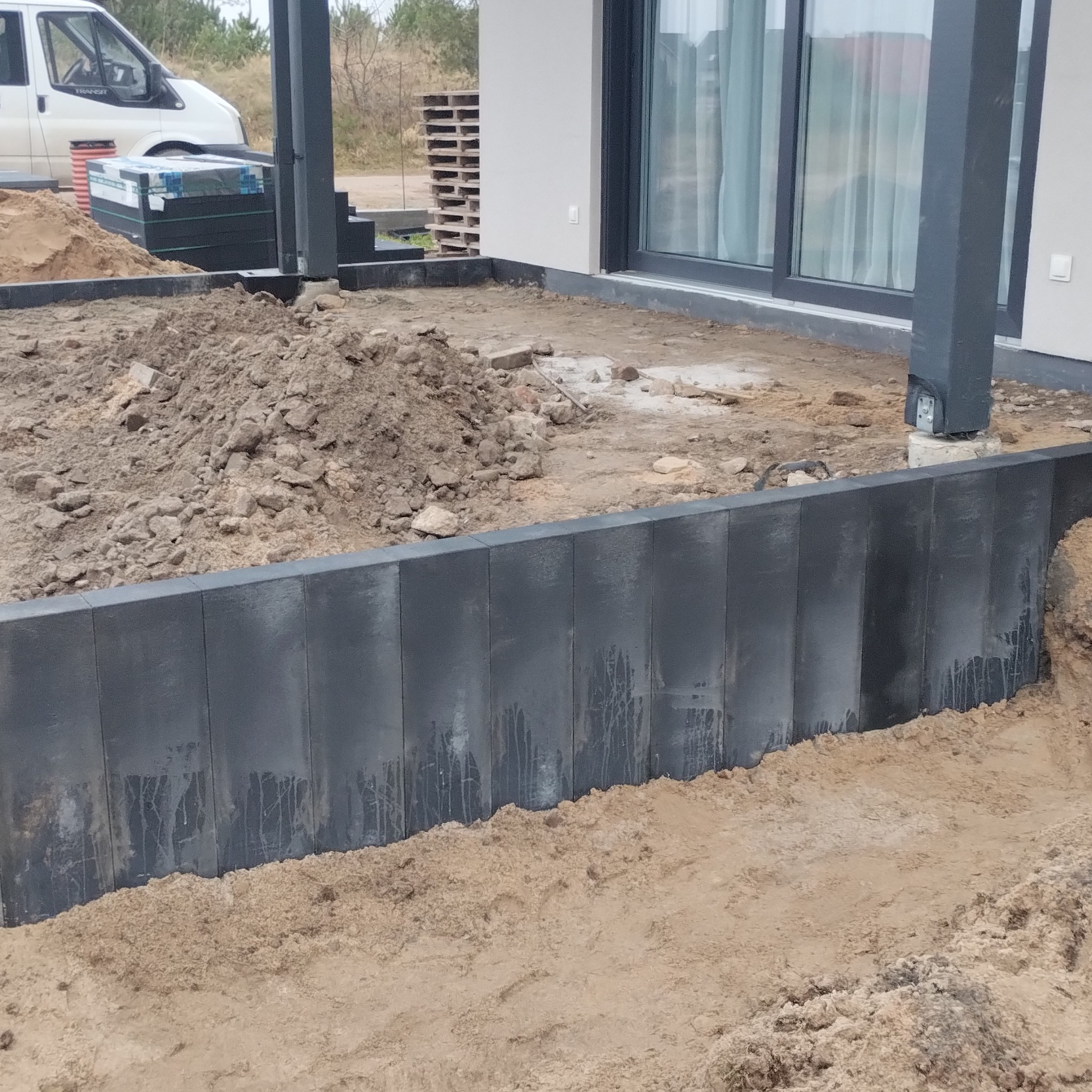 Ciemnoszara, pionowa palisada z betonu architektonicznego, częściowo obsypana piaskiem, tworząca obrzeże tarasu. W tle zaparkowany bus i palety.