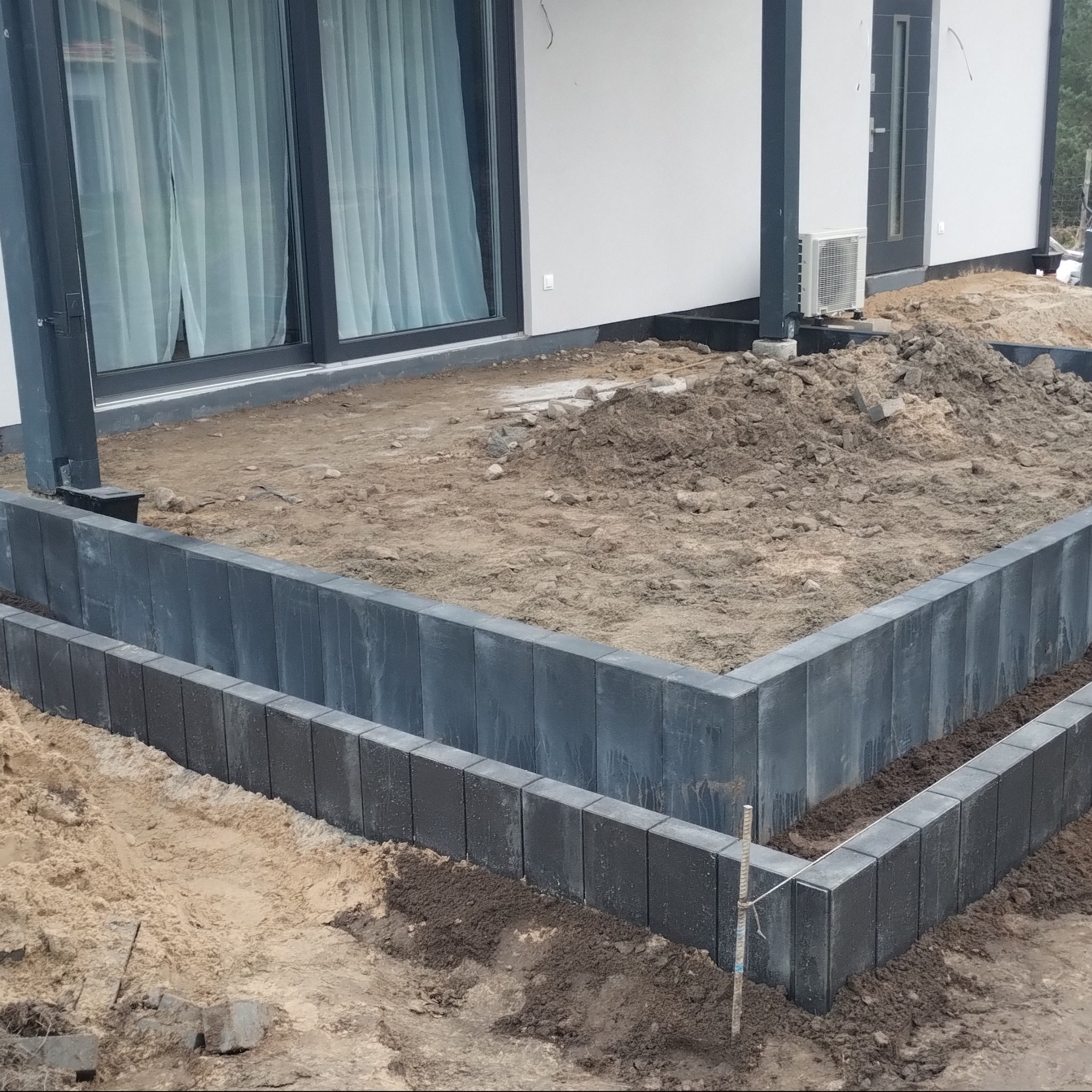 Etap budowy obrzeża z szarej kostki przy domu z oknami. Widoczny grunt i przygotowany teren pod dalsze prace brukarskie. Ujęcie z bliska, z widoczną fakturą kostki.