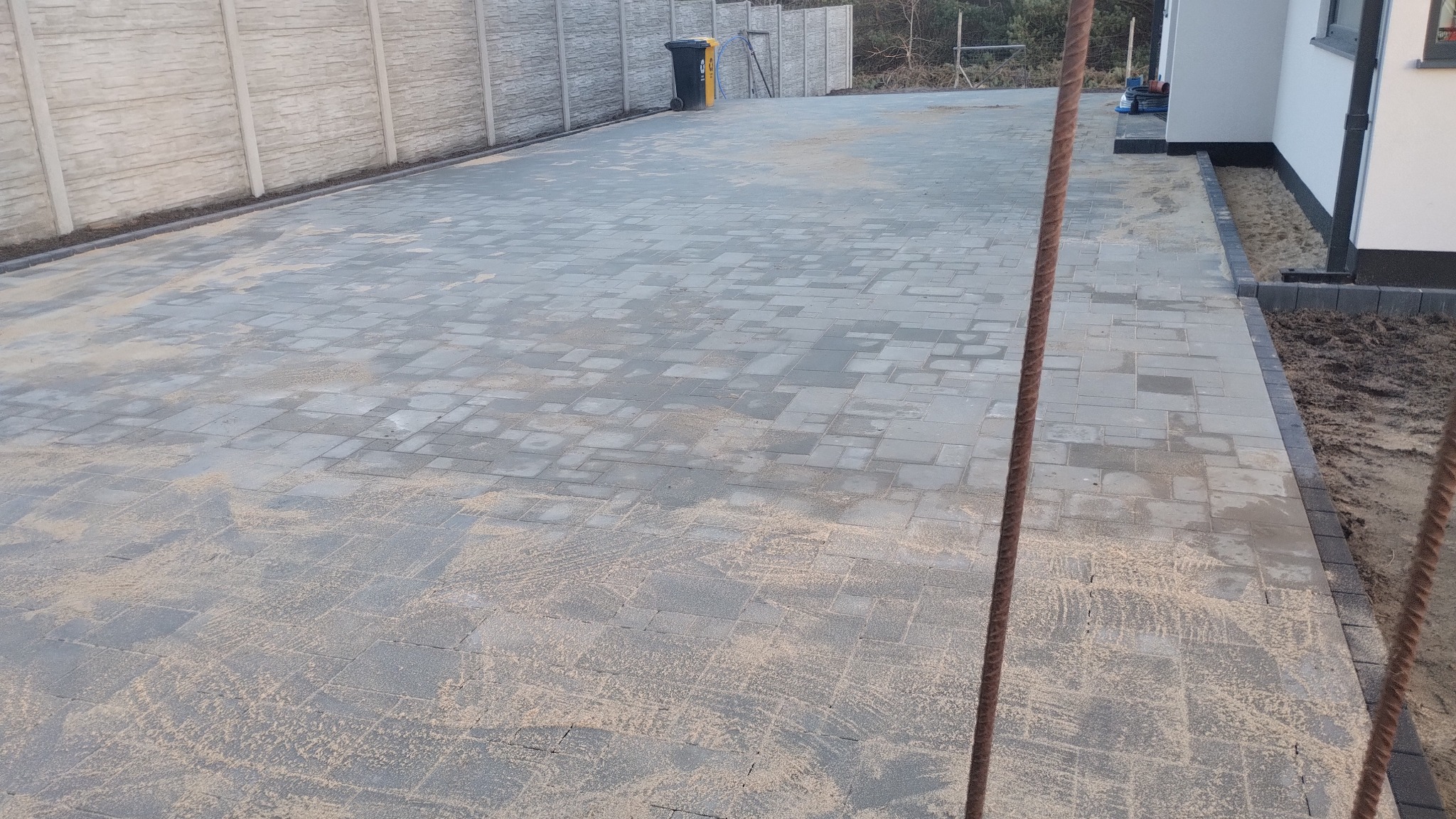 Szara kostka brukowa ułożona na podjeździe, widok z góry, z elementami ogrodzenia i fragmentem budynku w tle. Widoczne ślady piasku na powierzchni kostki.