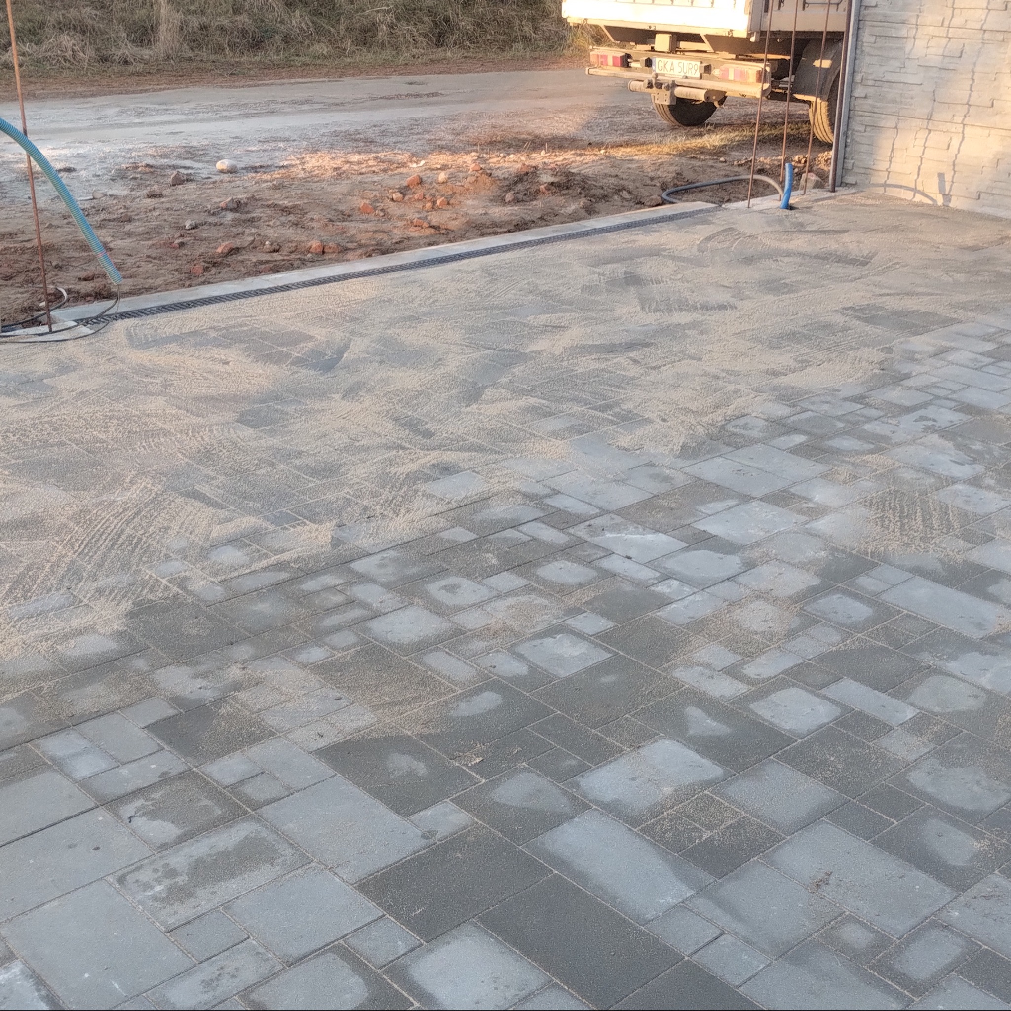 Ułożona, szara kostka brukowa z piaskiem na spoinach, widoczny fragment ciężarówki i obrzeże. Ujęcie z góry, w tle trawa i droga.