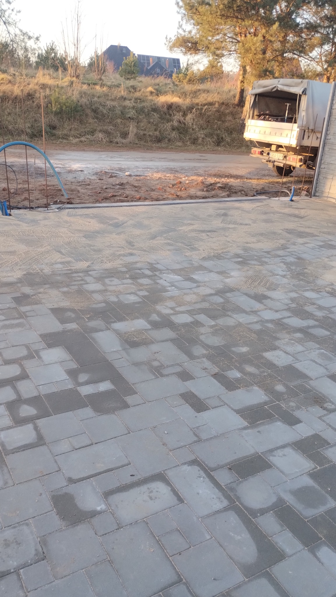 Ułożona, szara kostka brukowa o nieregularnym wzorze, z widocznym piaskiem w spoinach. W tle teren budowy, ciężarówka i dom z panelami słonecznymi.