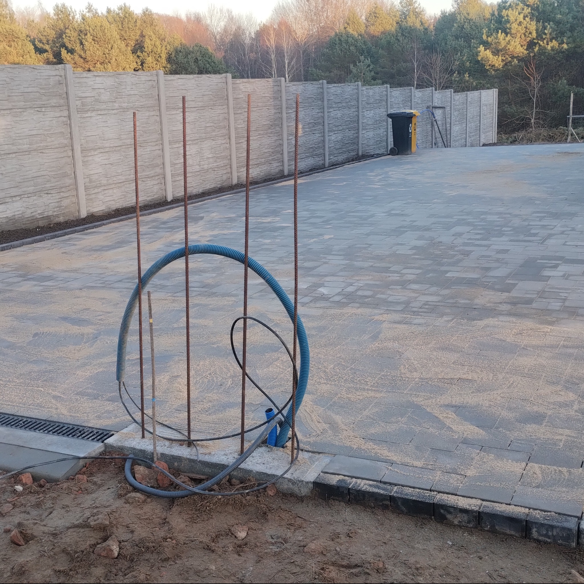 Układanie kostki brukowej w trakcie prac: widoczne kable w peszlu, wystające pręty zbrojeniowe i fragment ułożonej nawierzchni z kostki betonowej. W tle betonowe ogrodzenie.