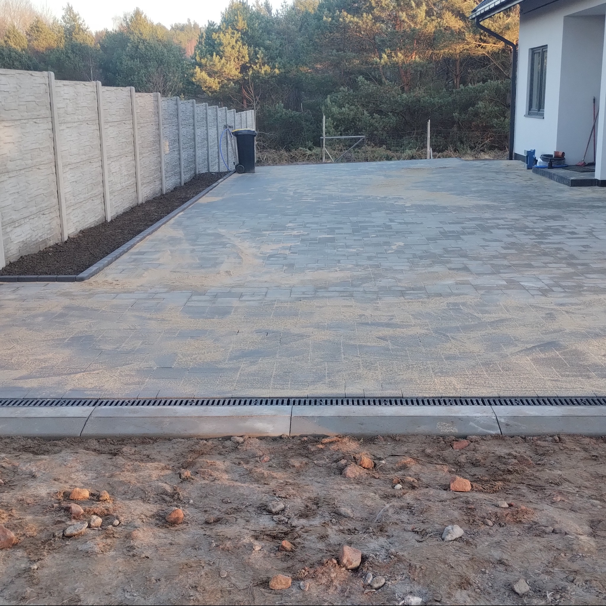 Ułożona kostka brukowa na podjeździe domu jednorodzinnego, widoczny drenaż liniowy, obrzeża i ogrodzenie panelowe. Tło stanowi las.
