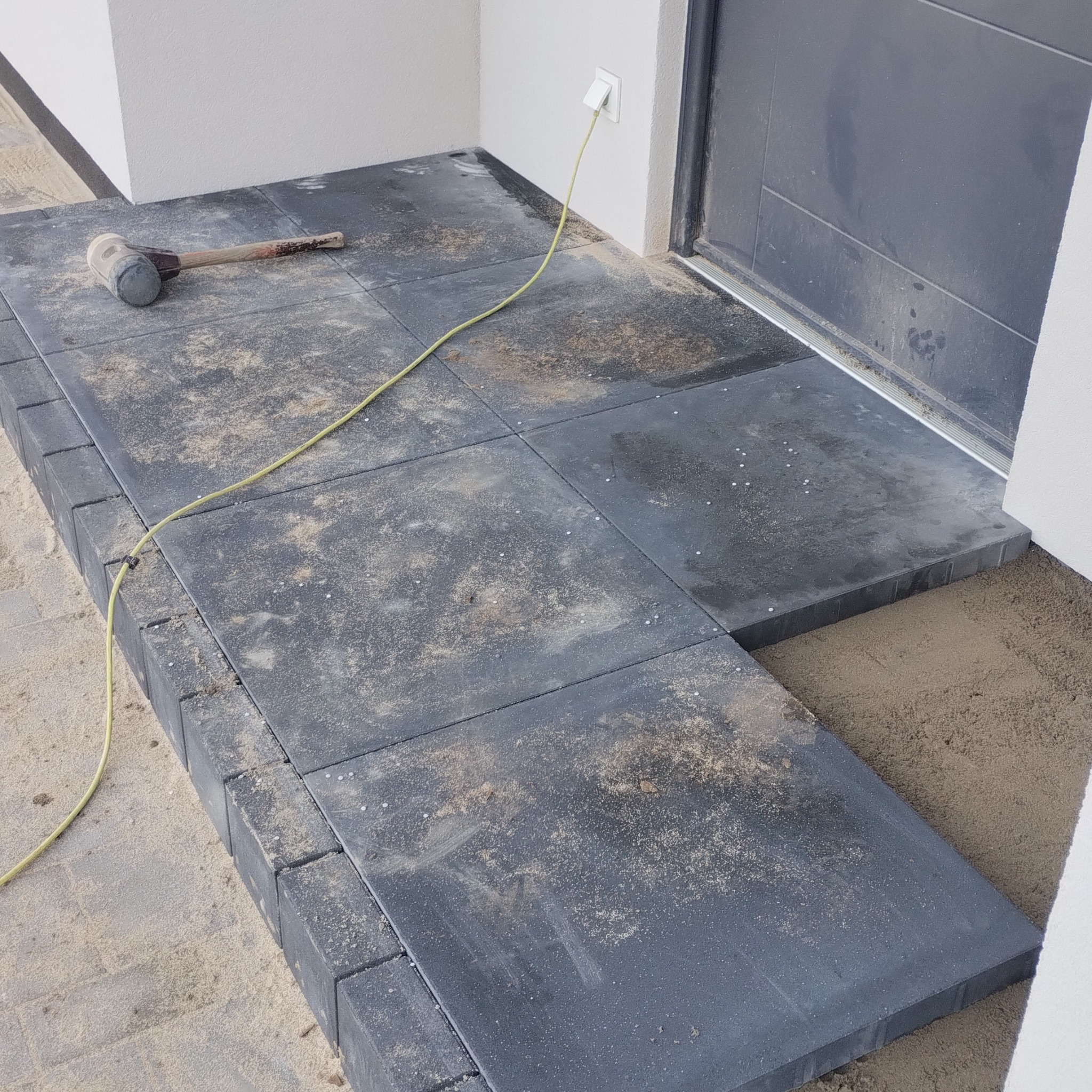 Ciemnoszara kostka brukowa ułożona przy wejściu, widoczny gumowy młotek i kabel zasilający. Piasek między kostkami. Częściowo ukończona praca.