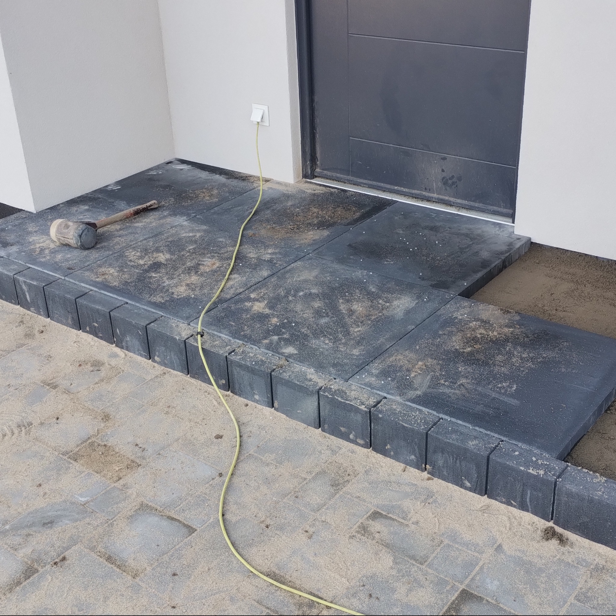 Ułożona, ciemnoszara kostka brukowa przed wejściem do domu, z widocznym gumowym młotkiem i kablem zasilającym. Krawężniki z kostki. Piasek na powierzchni.