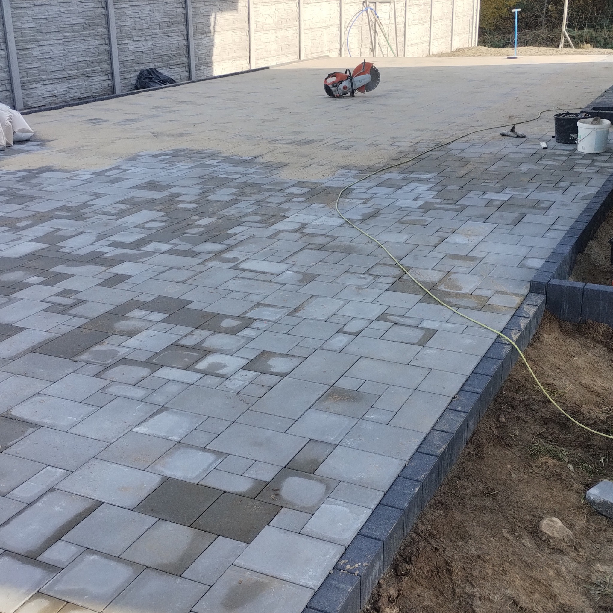 Ułożona, szara kostka brukowa w nieregularny wzór, obrzeża z ciemnej kostki. W tle piła do cięcia kostki i narzędzia. Częściowo mokra nawierzchnia.