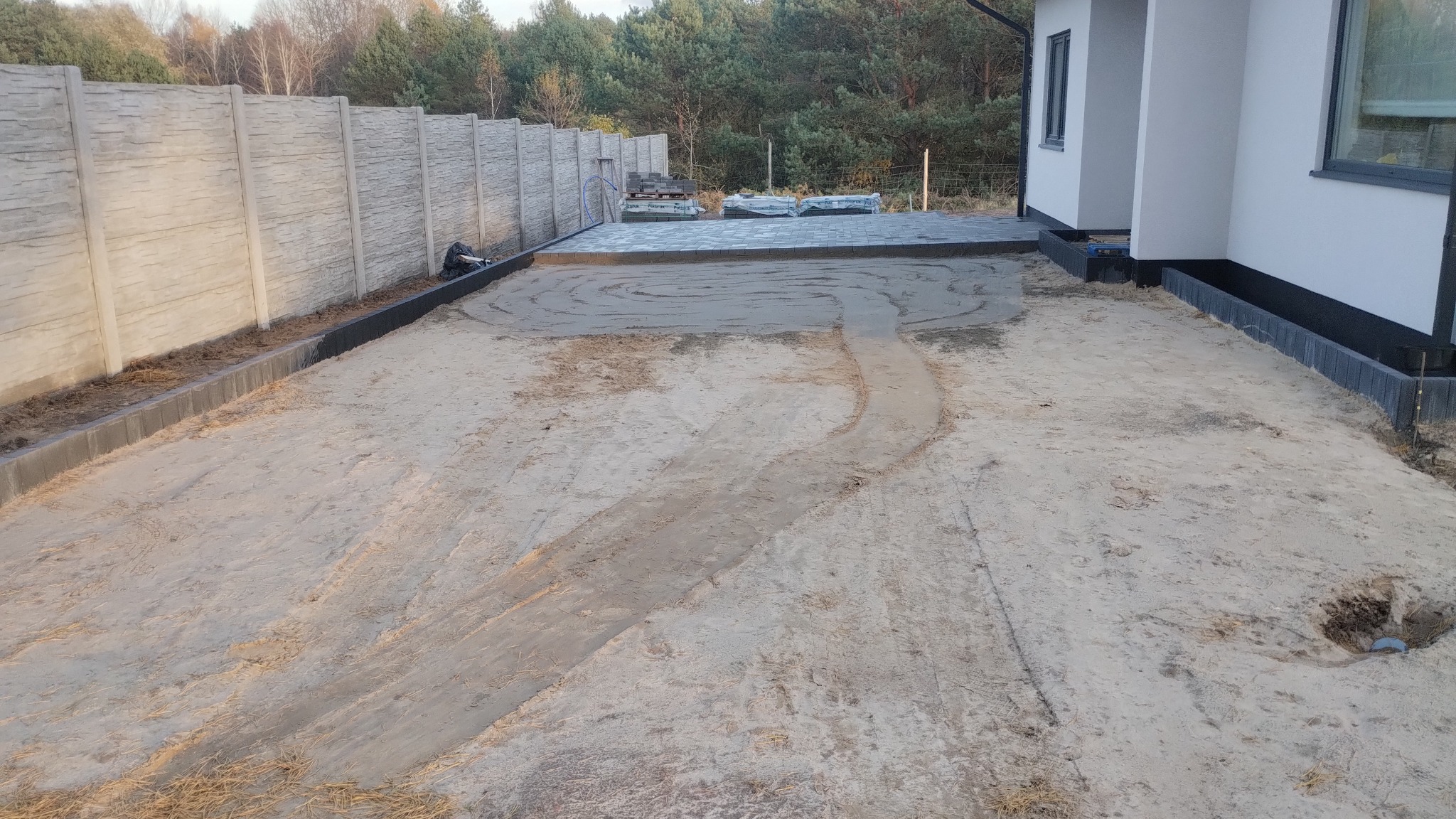 Etap prac: przygotowanie podłoża pod kostkę brukową. Widoczne obrzeża, podsypka piaskowa i częściowo ułożona kostka przy budynku. Tło: betonowy płot i drzewa.