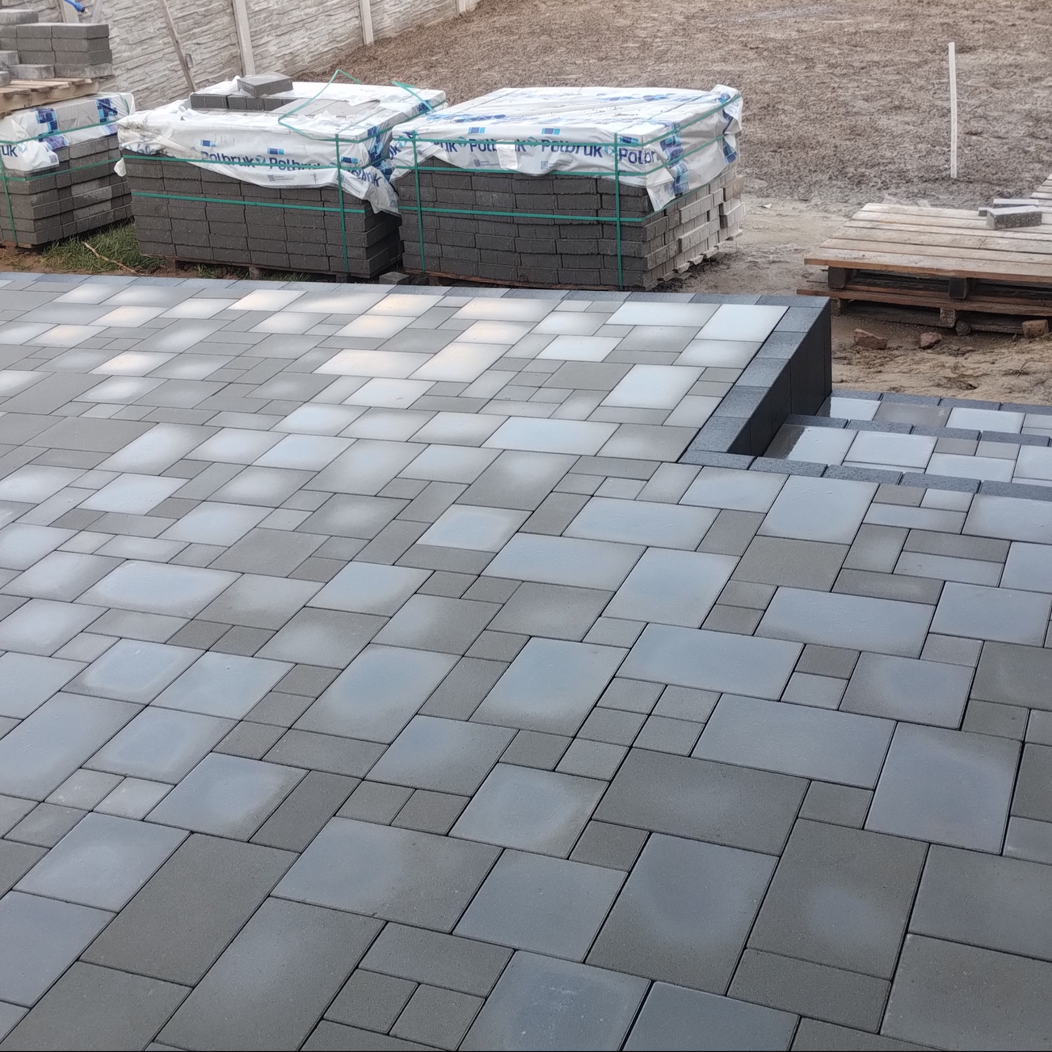 Ułożona, szara kostka brukowa tworząca wzór na podjeździe, z paletami kostki w tle. Widoczne schody z kostki brukowej. Całość w stonowanej kolorystyce.