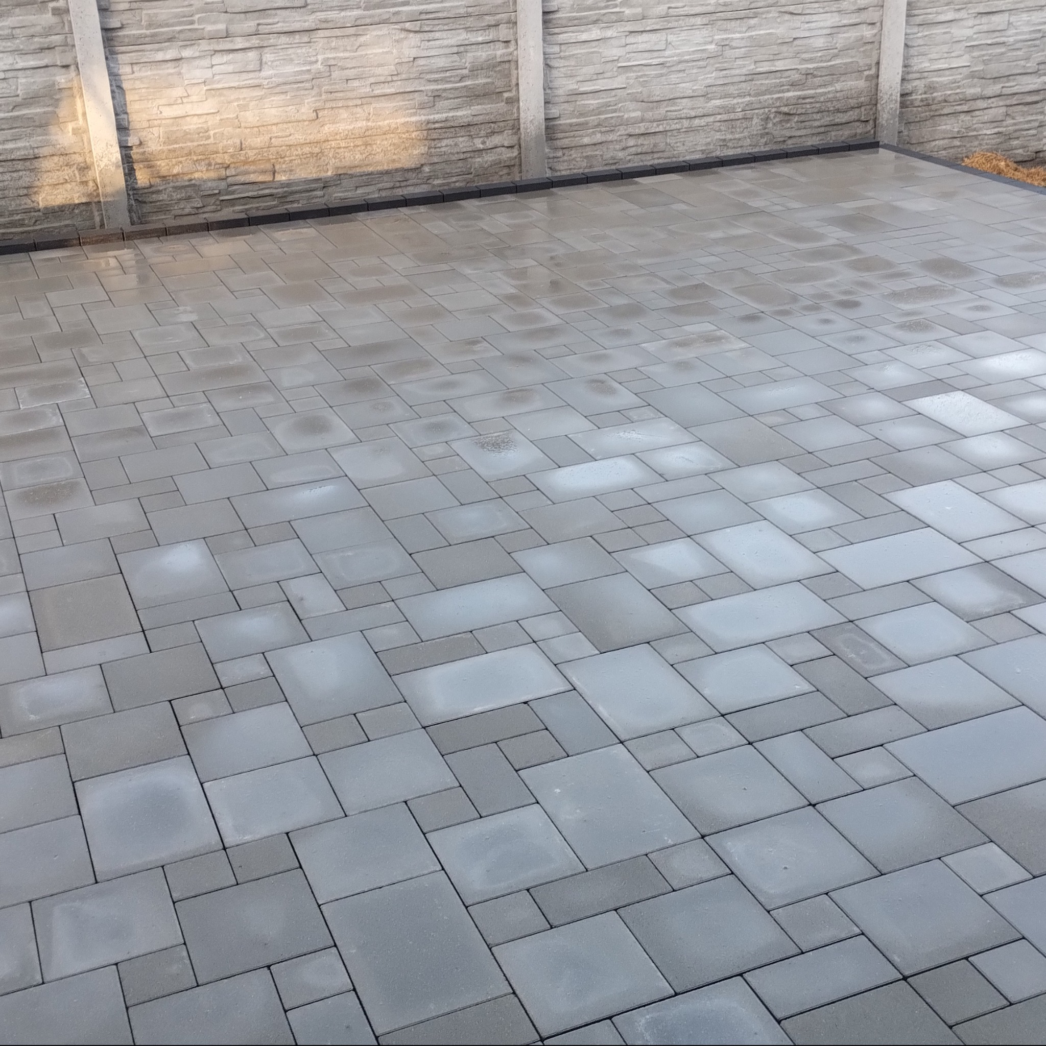 Szara kostka brukowa ułożona w geometryczny wzór, otoczona betonowym ogrodzeniem. Widok z góry ukazuje regularność i precyzję wykonania.