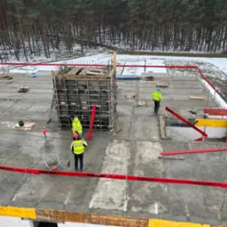Betonowy fundament budynku w trakcie budowy, widoczne szalunki, zbrojenia i pracownicy w odblaskowych kamizelkach. W tle las i częściowo ośnieżony teren.