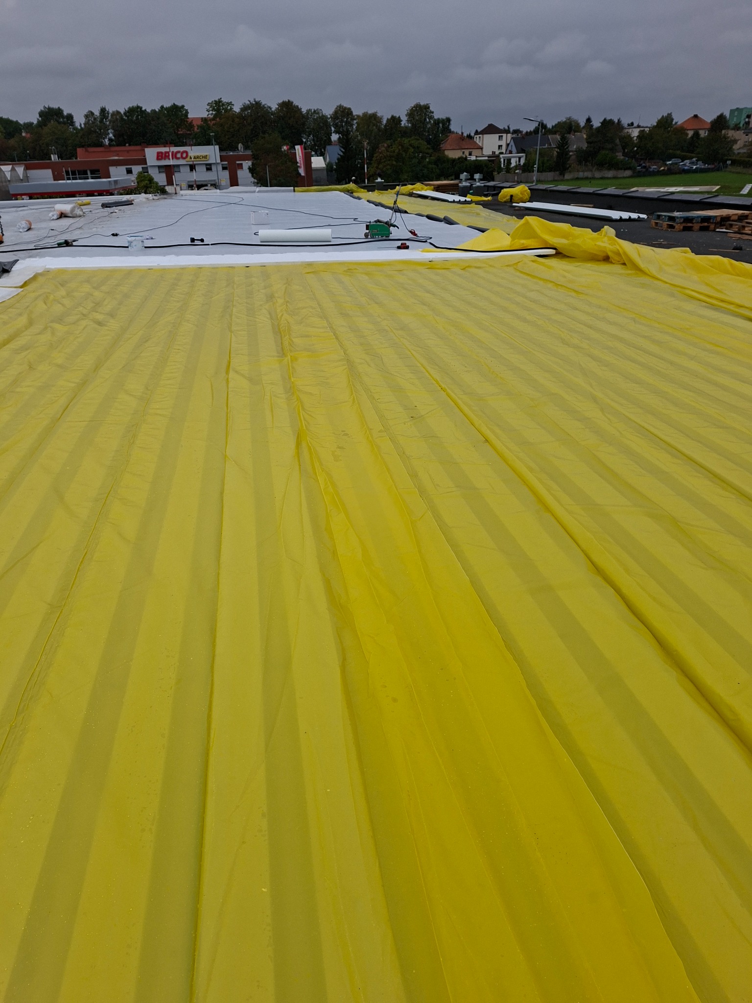 Żółta membrana dachowa rozłożona na płaskim dachu budynku, widoczne narzędzia i materiały izolacyjne, budynek Brico Marche w tle.