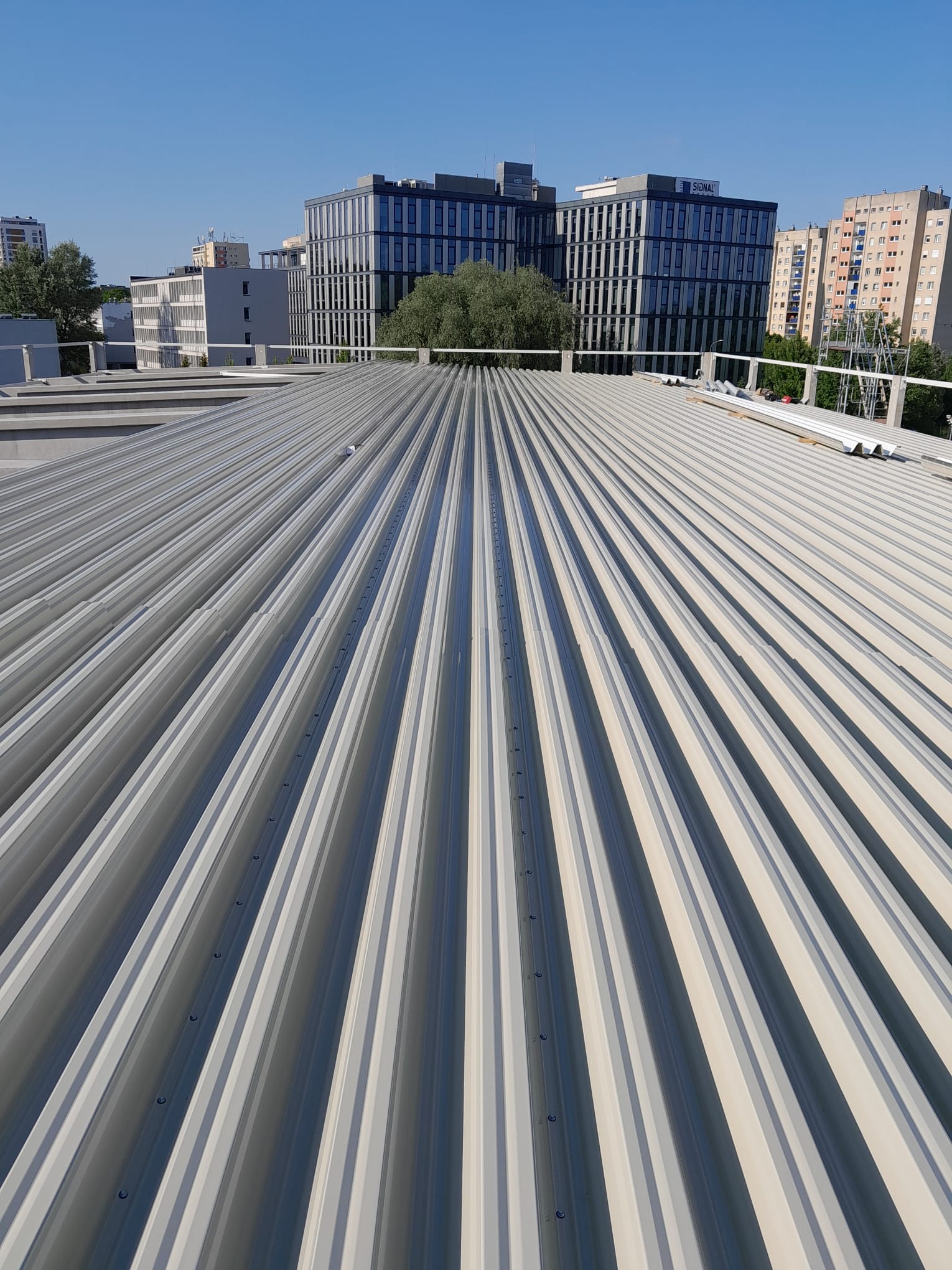 Nowoczesny, blaszany dach trapezowy w kolorze jasnoszarym, z widocznymi mocowaniami, na tle miejskiego krajobrazu z budynkami biurowymi i błękitnym niebem.