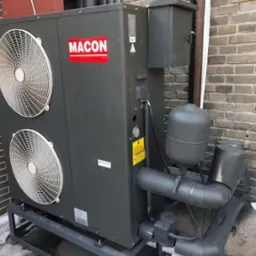 Szara pompa ciepła z dwoma wentylatorami, zamontowana na metalowej podstawie przy ceglanej ścianie. Widoczne rury i zbiornik wyrównawczy.