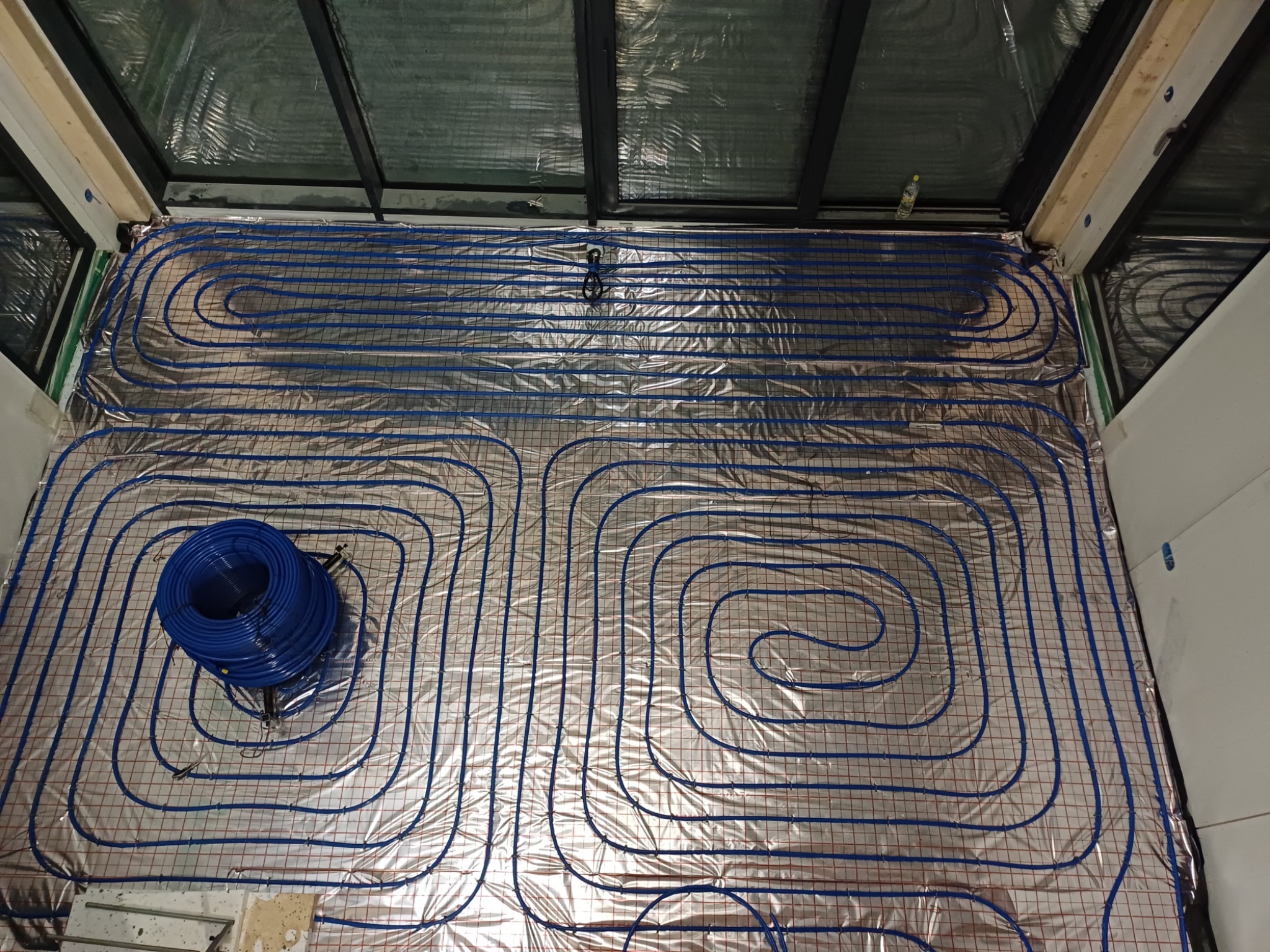 Widok z góry na rozłożone rury ogrzewania podłogowego w pomieszczeniu z przeszklonym dachem, widoczna rolka niebieskich rur i folia izolacyjna z siatką.