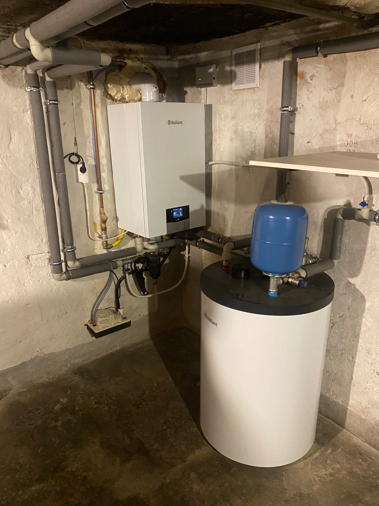 Biały piec grzewczy Vaillant zamontowany na ścianie w piwnicy, z podłączonymi rurami, zbiornikiem wyrównawczym i pompą skroplin.