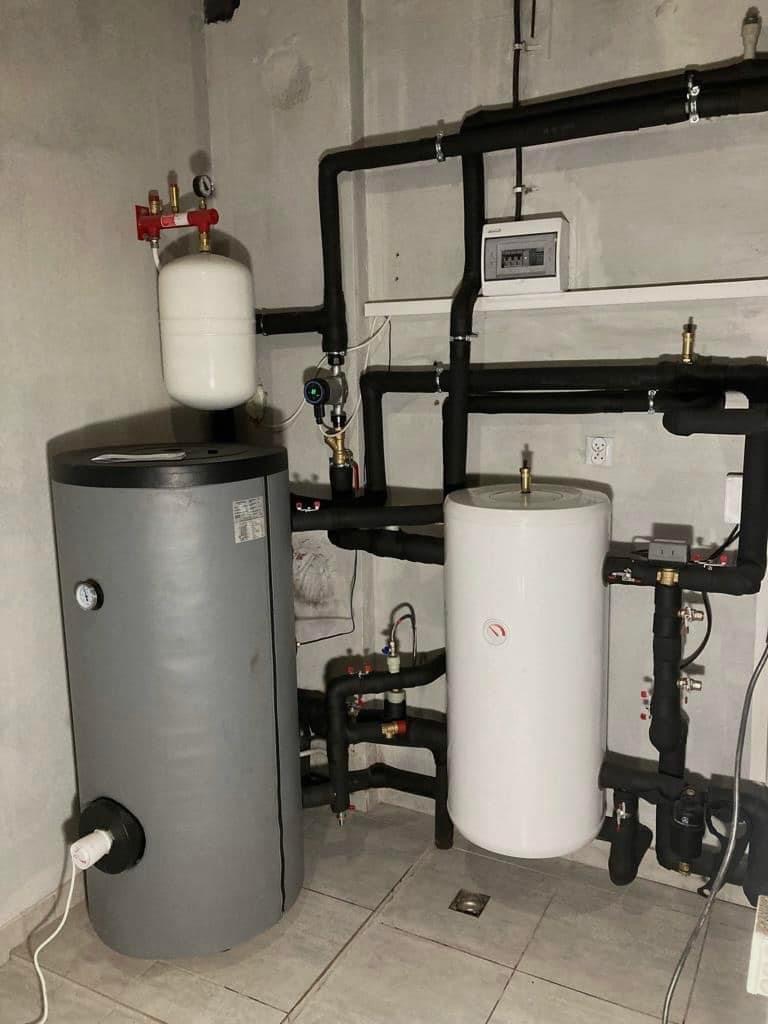 Szary i biały zbiornik buforowy w kotłowni z czarnymi rurami instalacji grzewczej, widoczna skrzynka elektryczna na ścianie i płytki podłogowe.