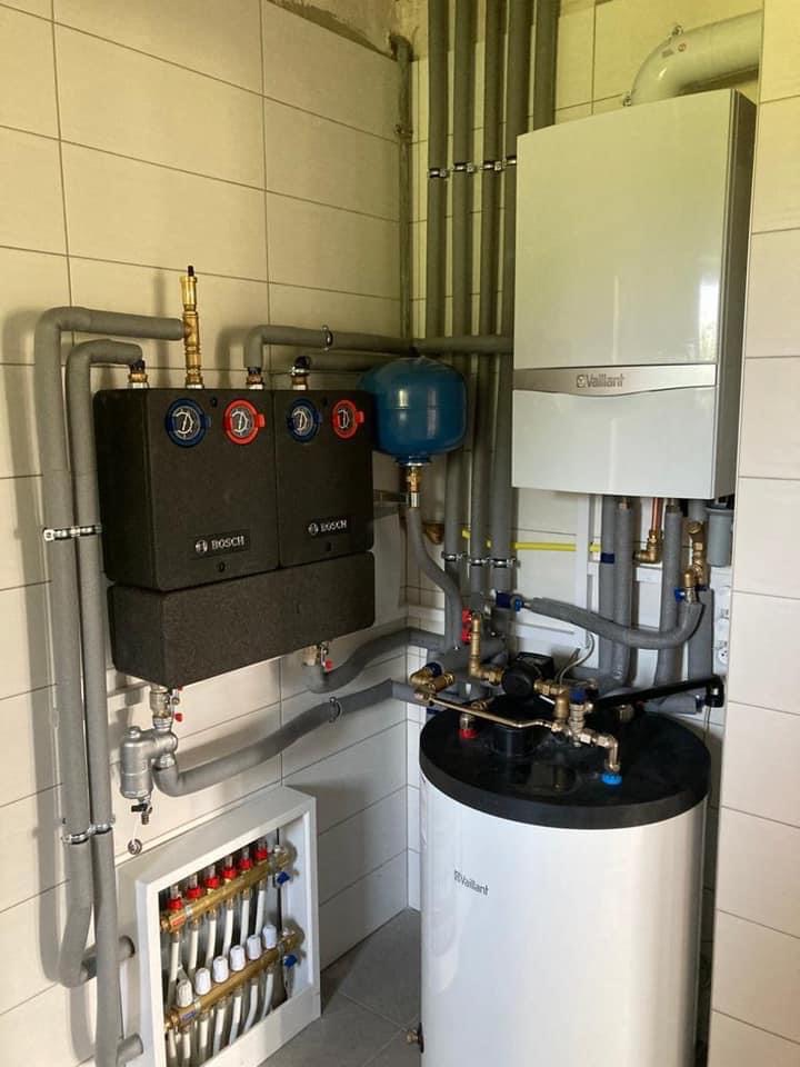 Kompletna instalacja grzewcza w kotłowni: dwa moduły Bosch, biały zasobnik i piec Vaillant, liczne rury i zawory, rozdzielacz ogrzewania podłogowego w szafce na ścianie z białych płytek.