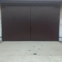 Zamknięta, dwuskrzydłowa brama garażowa w kolorze ciemnego brązu, z widocznymi pionowymi przetłoczeniami, zamontowana w budynku z białych bloczków.