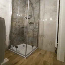 Wykończona kabina prysznicowa z przeszklonymi drzwiami, zamontowana na białym brodziku, z widoczną armaturą i płytkami imitującymi kamień na ścianach oraz drewnopodobną podłogą.