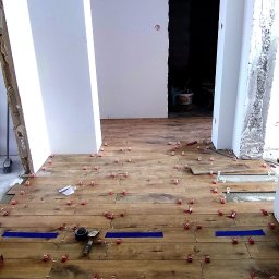 Kompleksowe wykończenia wnętrz - Układanie płytek imitujących drewno, widoczne poziomice, krzyżyki dystansowe, niebieska taśma maskująca, pomieszczenie w trakcie prac wykończeniowych.