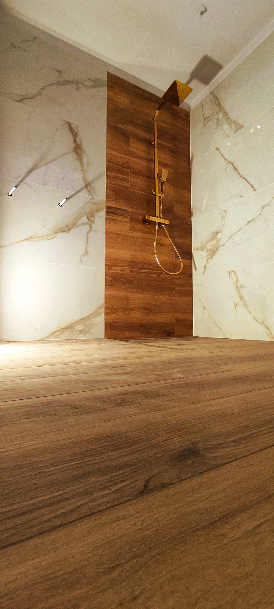 Nowoczesna łazienka z drewnopodobną podłogą i ścianą prysznicową, białe marmurowe płytki na pozostałych ścianach, złota armatura prysznicowa z deszczownicą.