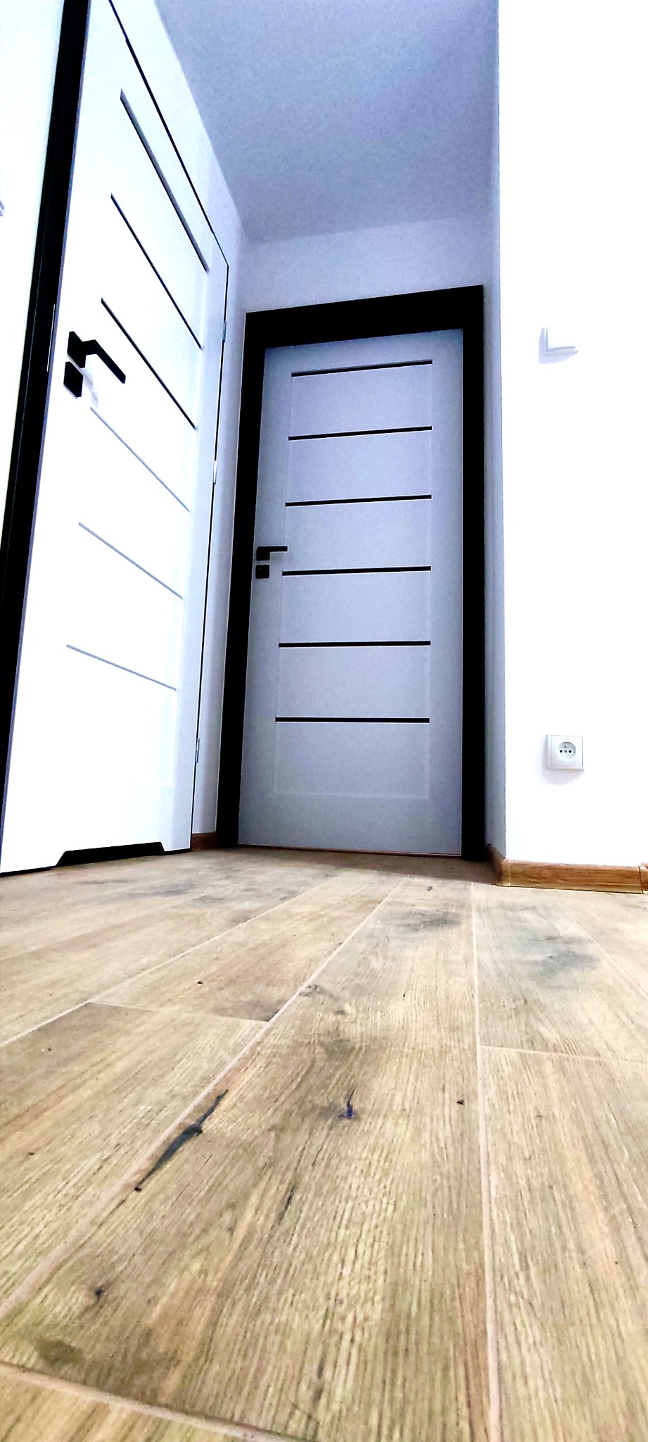 Nowoczesne, białe drzwi wewnętrzne z czarnymi akcentami i drewnopodobna podłoga ułożona w jodełkę, widok z niskiej perspektywy.
